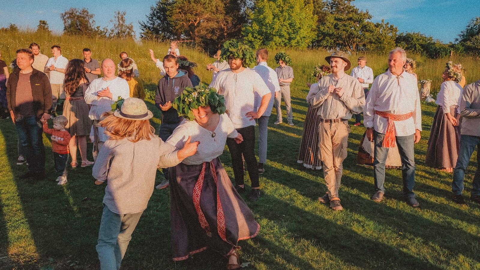 People gathered in a sunlit meadow at a midsummer celebration, several wearing large oak-leaf and flower wreaths and folk dress with woven sashes, while a woman in a wreath dances with a companion in the foreground