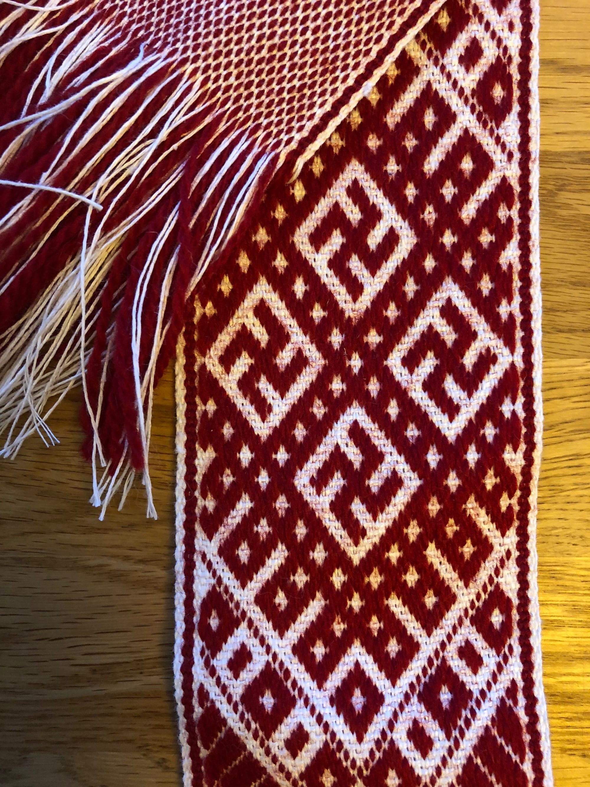 A close view of a red and white woven belt with a dense pattern of interlocking geometric diamonds, its fringed end lying across a wooden surface