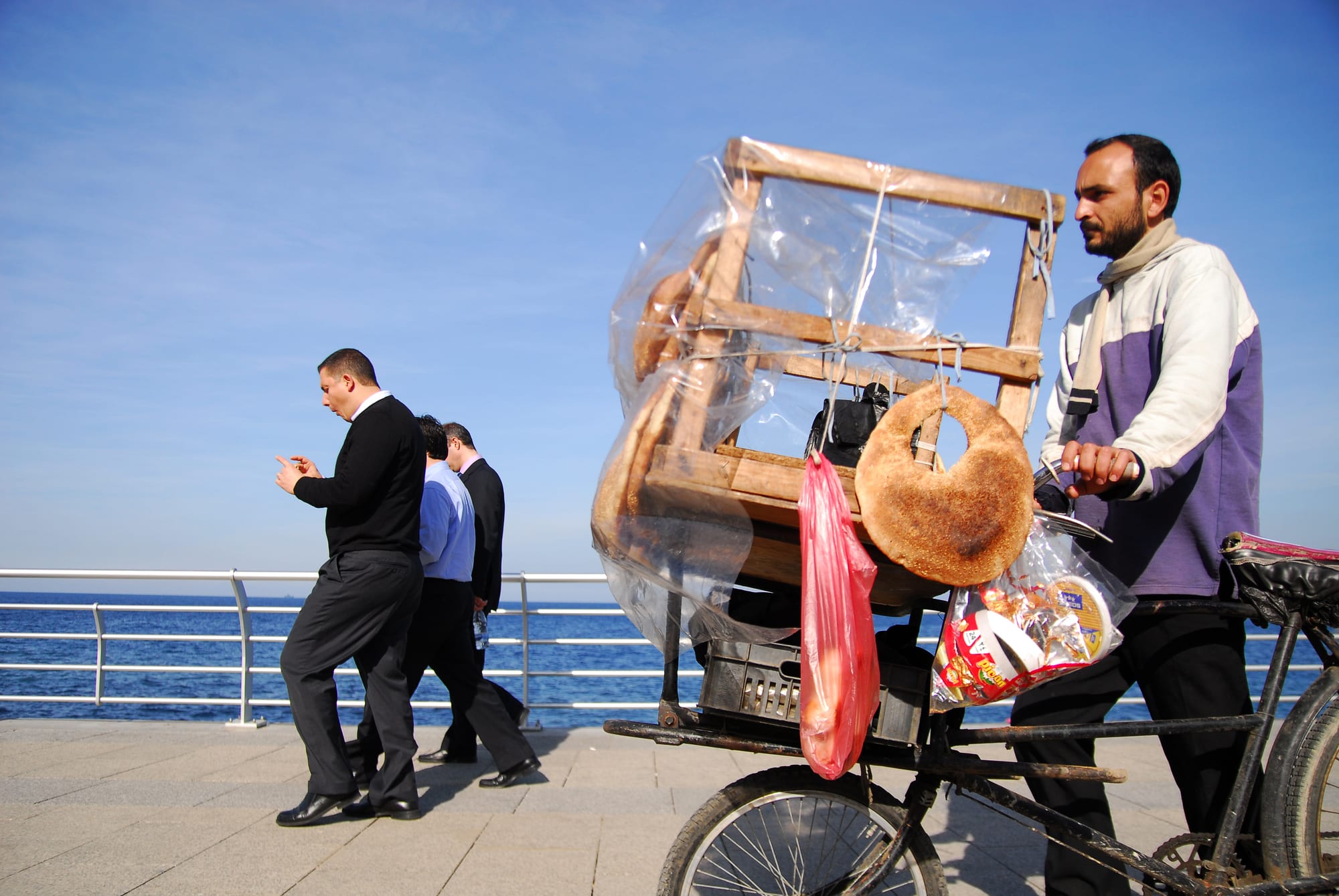 A street vendor pushing a bicycle loaded with rings of sesame bread along Beirut's seafront promenade, with pedestrians walking past and the sea behind