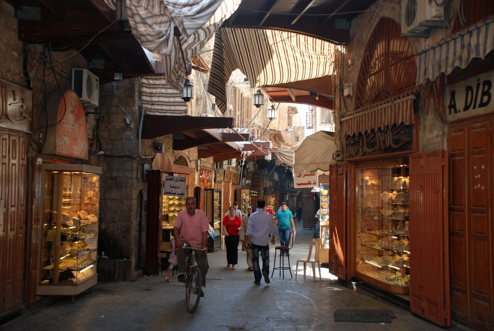 A narrow, covered market lane lined with gold and jewellery shops, with several people walking and a man cycling past