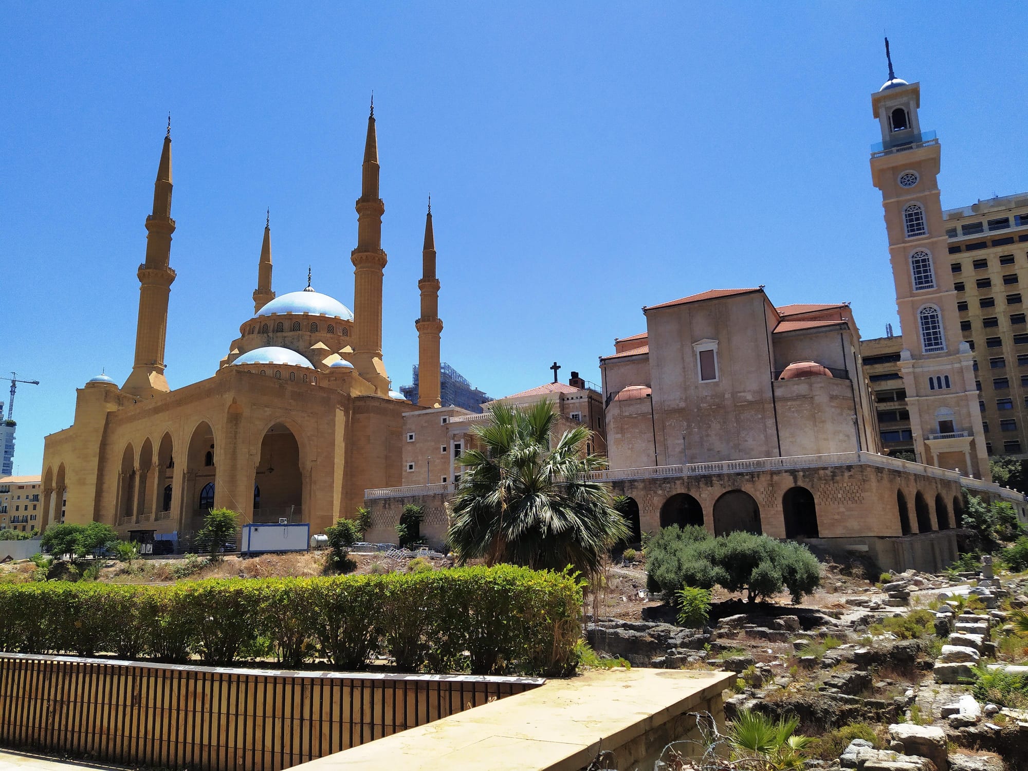 A large sandstone mosque with four slender minarets and blue domes stands beside a church with a tall clock tower topped by a cross, under a clear blue sky