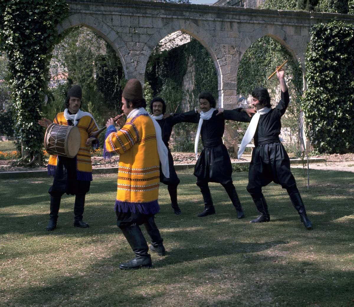 Male dancers in black costumes and white scarves with arms linked across each other's shoulders dance on a lawn in front of a stone arcade, accompanied by a drummer and a pipe player in bright yellow embroidered jackets