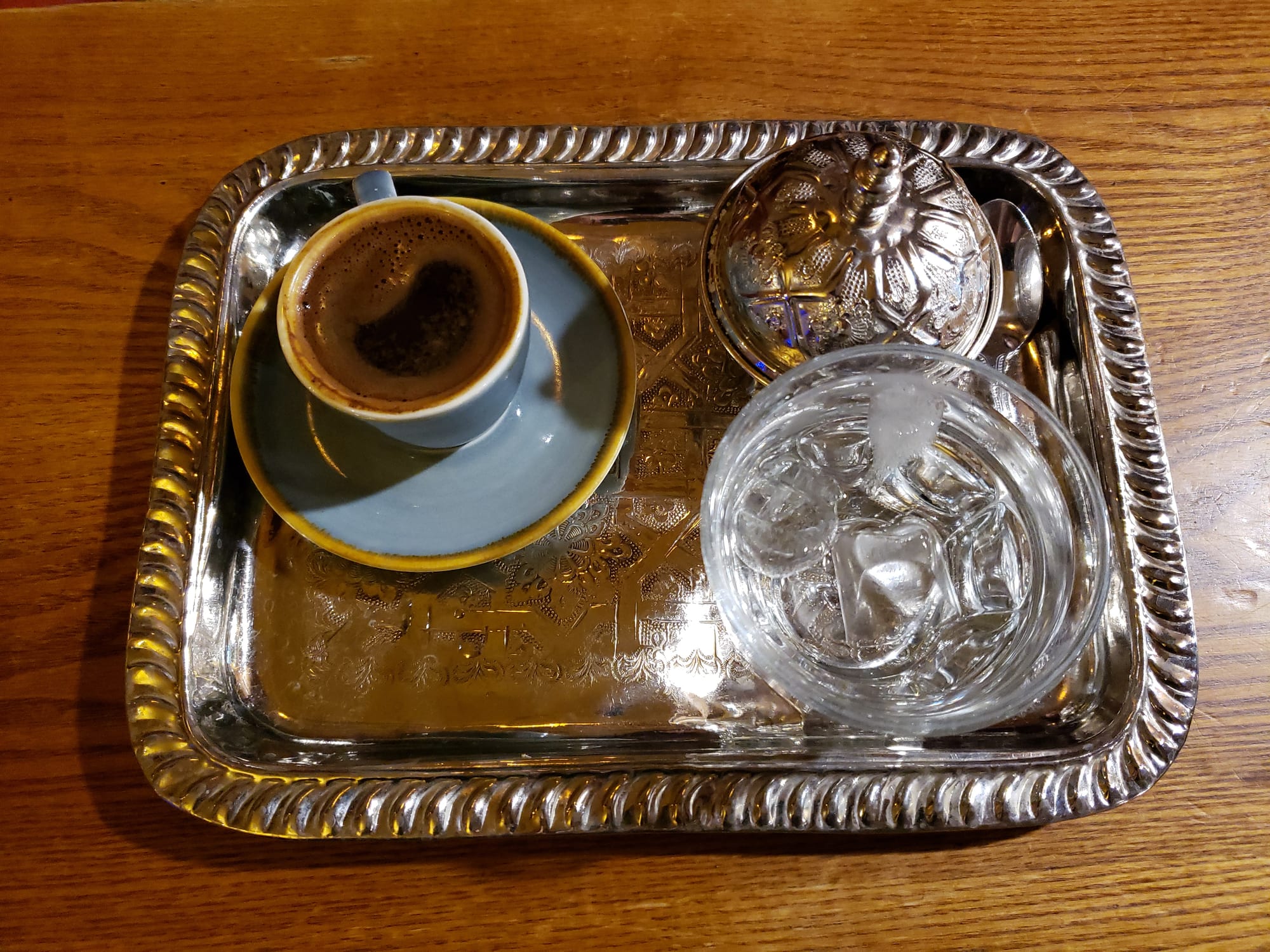 A small cup of dark coffee on a saucer, set on an engraved silver tray beside a glass of iced water and a silver lidded sugar bowl