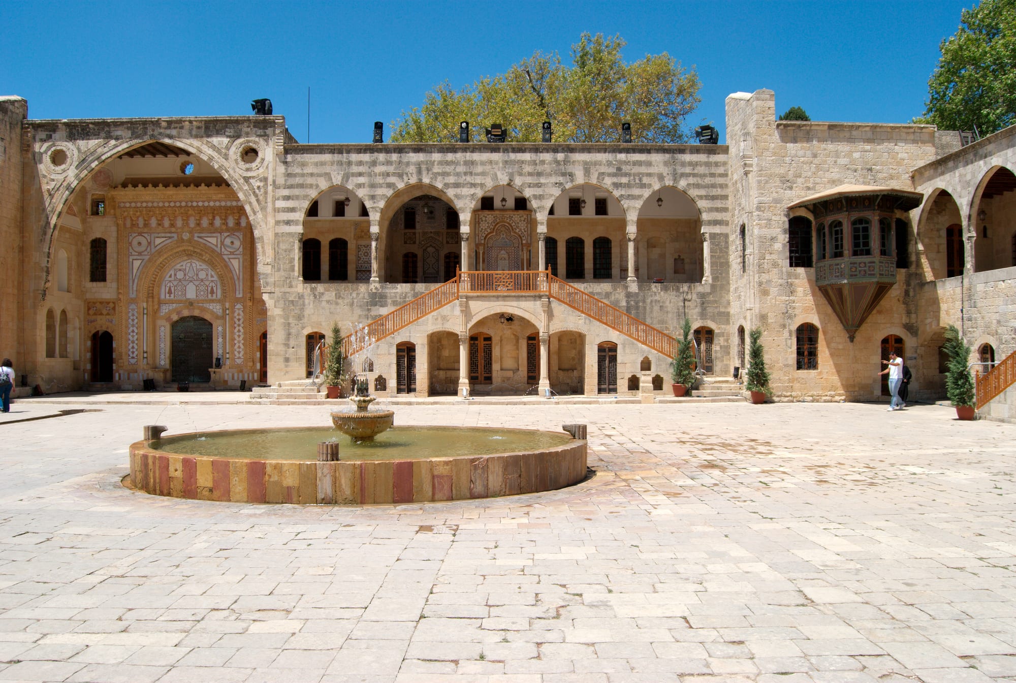 A wide stone-paved palace courtyard with a round fountain, ringed by arcaded galleries, a double staircase and an ornate inlaid entrance portal