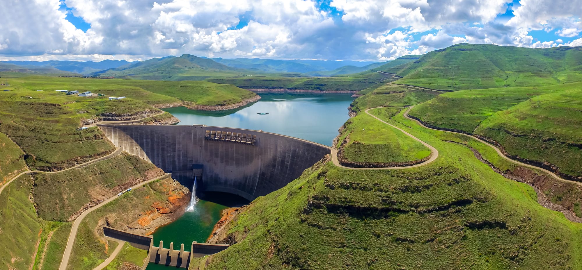 A tall curved concrete dam wall holding back a long reservoir in a mountain valley