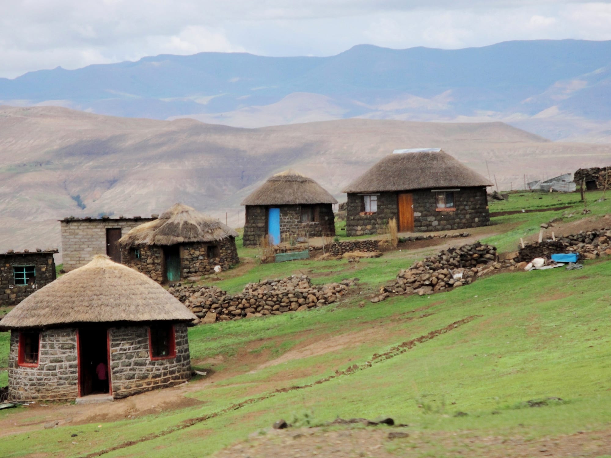 A cluster of round stone huts with thick thatched roofs on a highland hillside, with mountains and cloud in the distance