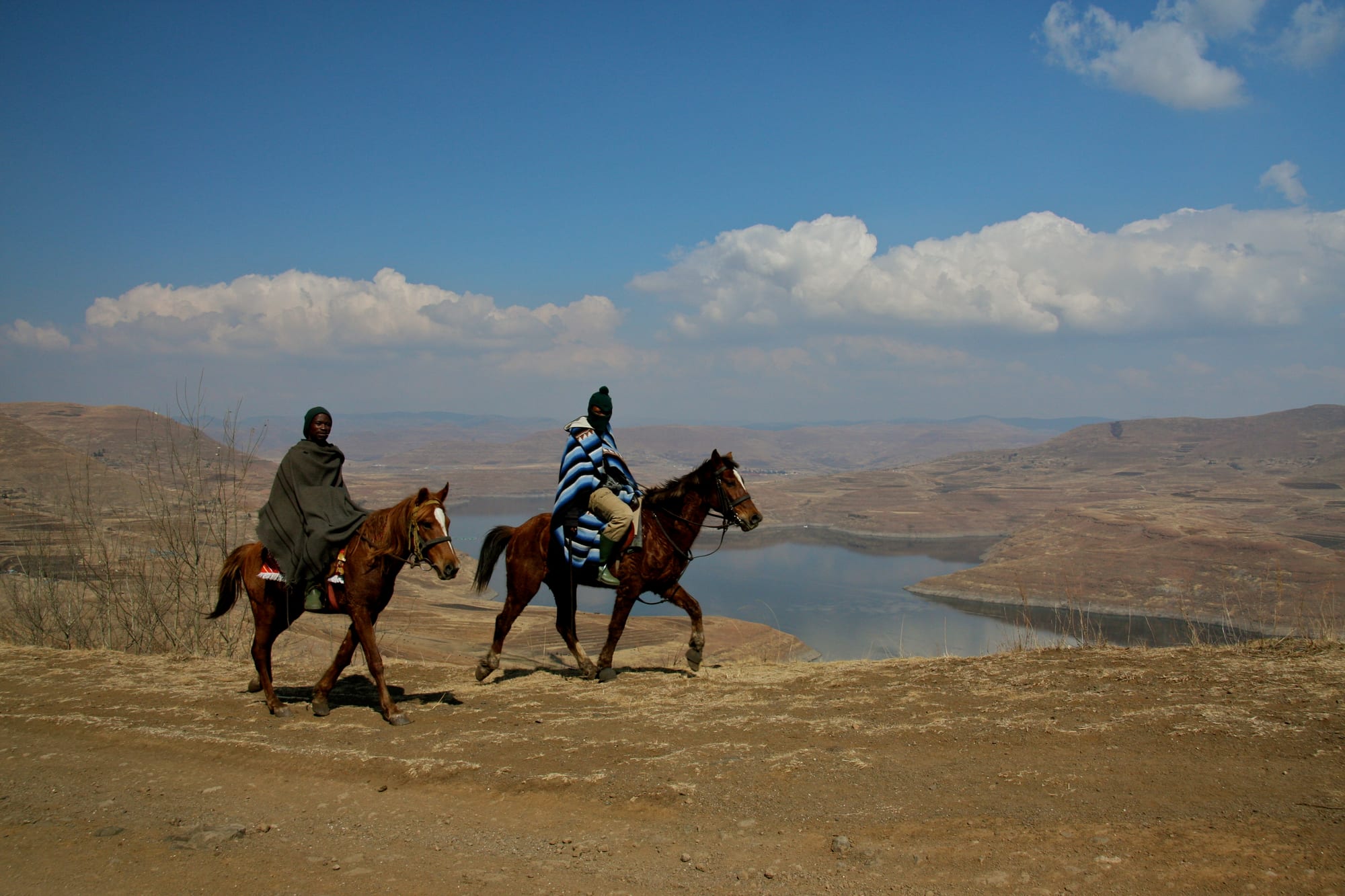 Two riders on brown horses cross a dry dirt track, one wrapped in a dark grey blanket and the other in a blue and white striped blanket, with a winding lake and bare brown hills behind under a blue sky