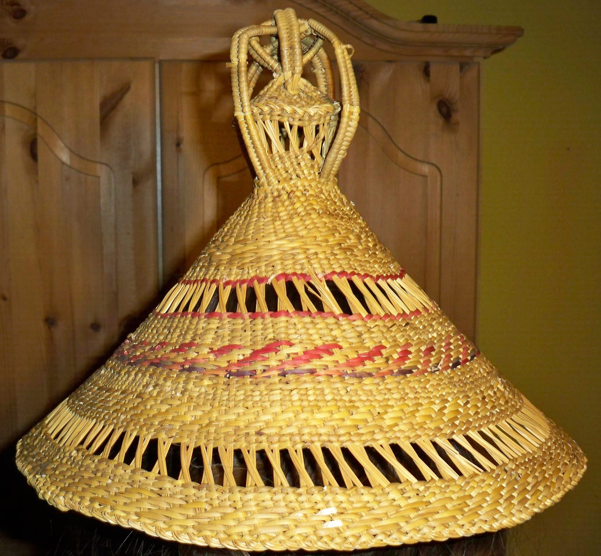 A conical woven straw hat with bands of red and purple fibre and open-work rows, topped by a knotted, loop-shaped crown
