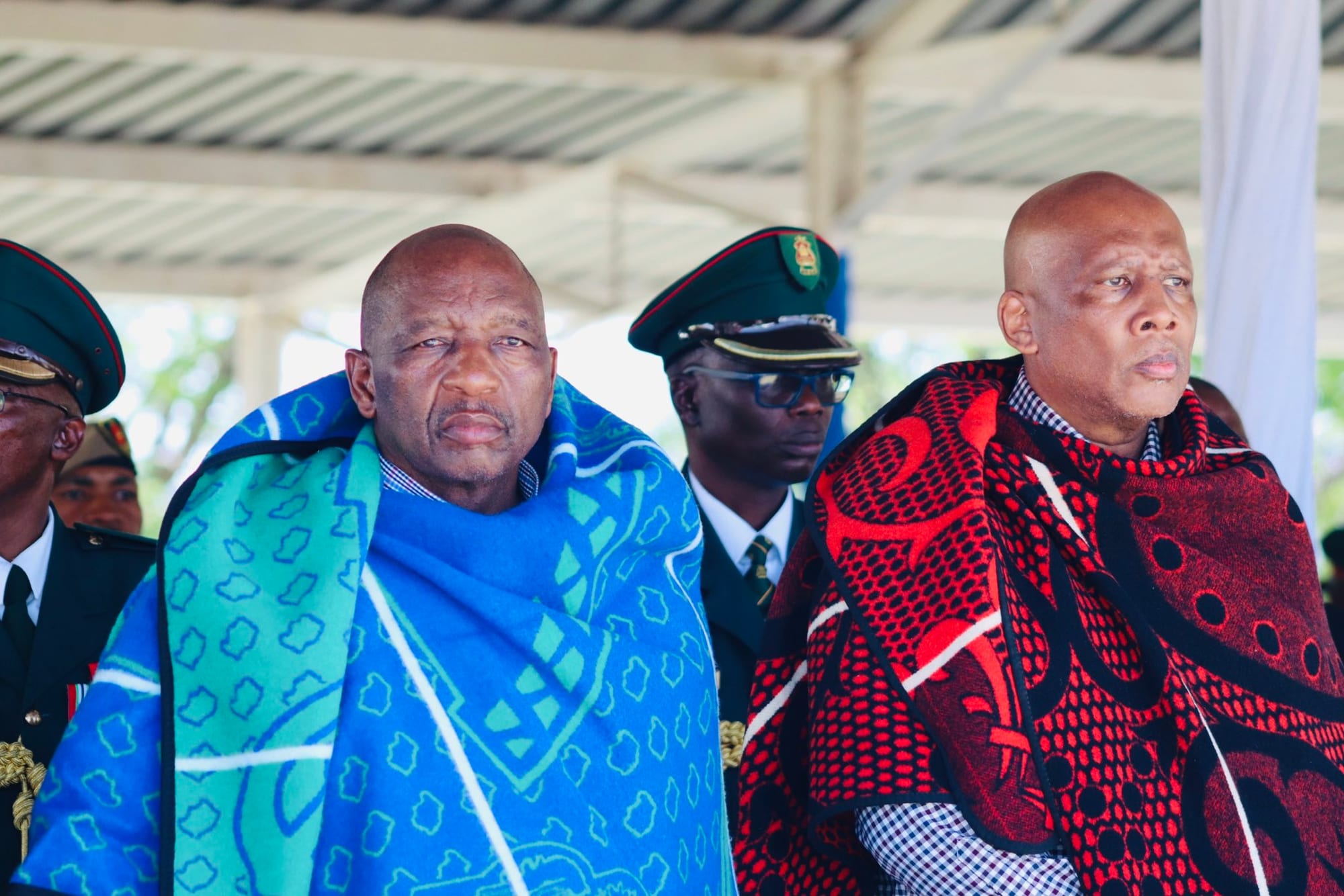 Two men seated under a canopy, each wrapped in a patterned Basotho blanket, one in blue and green and one in red and black, with uniformed guards in peaked caps standing behind them