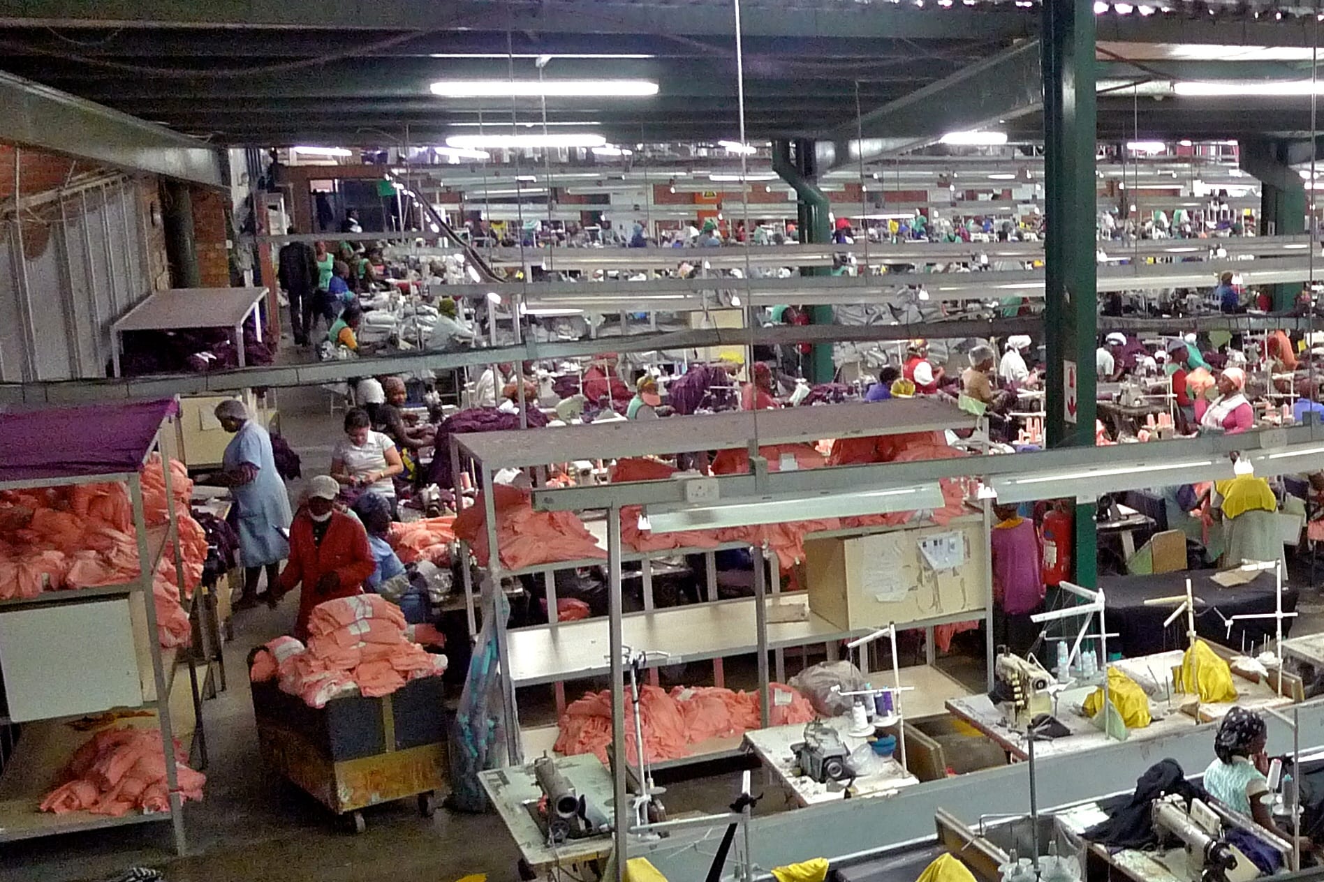A large factory floor seen from above, with rows of workers at sewing tables under strip lights and carts piled with pink garments