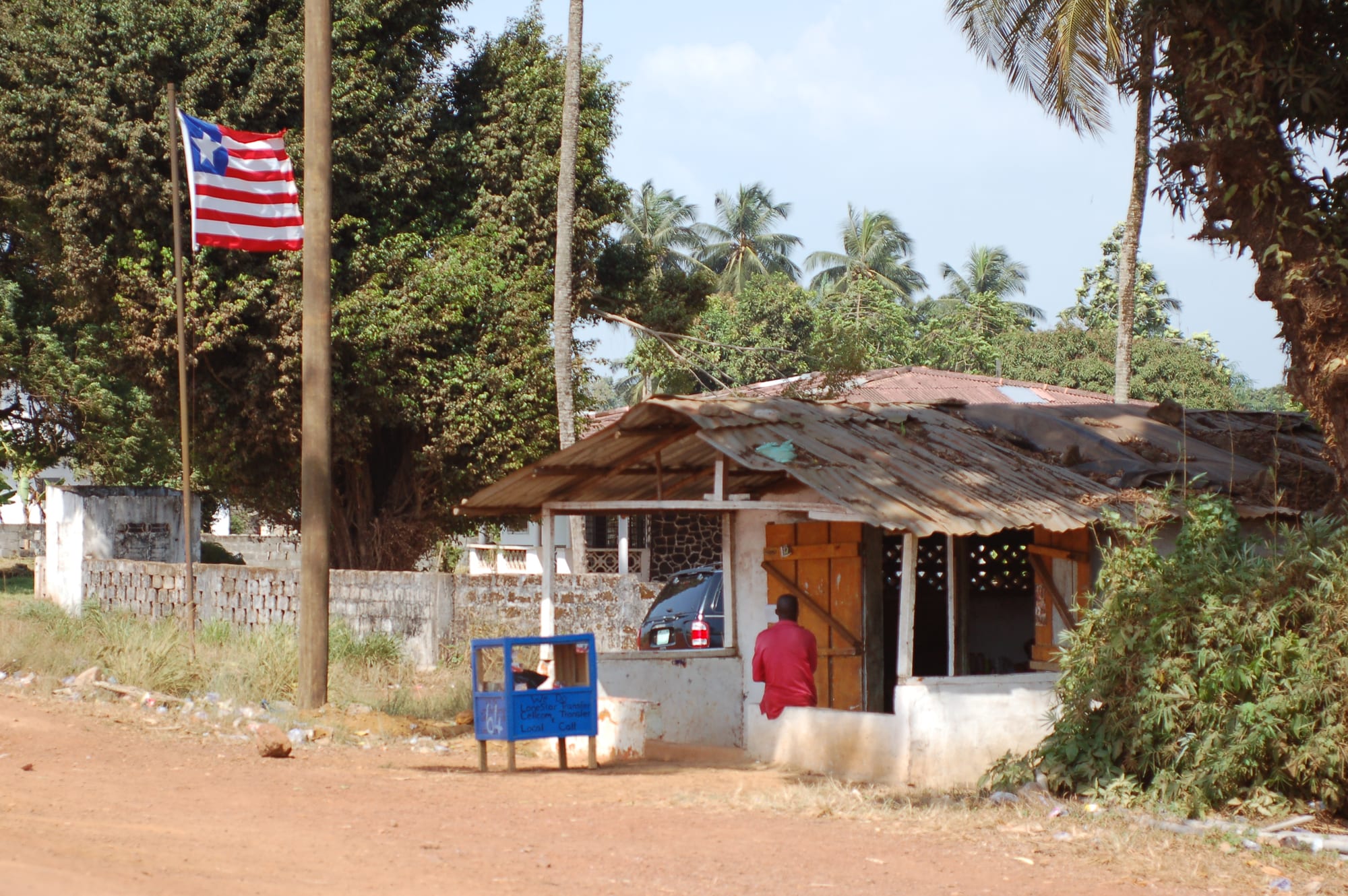 The red, white and blue Liberian flag with a single white star flies from a pole beside a small roadside shop with a corrugated-iron roof, with palm trees behind