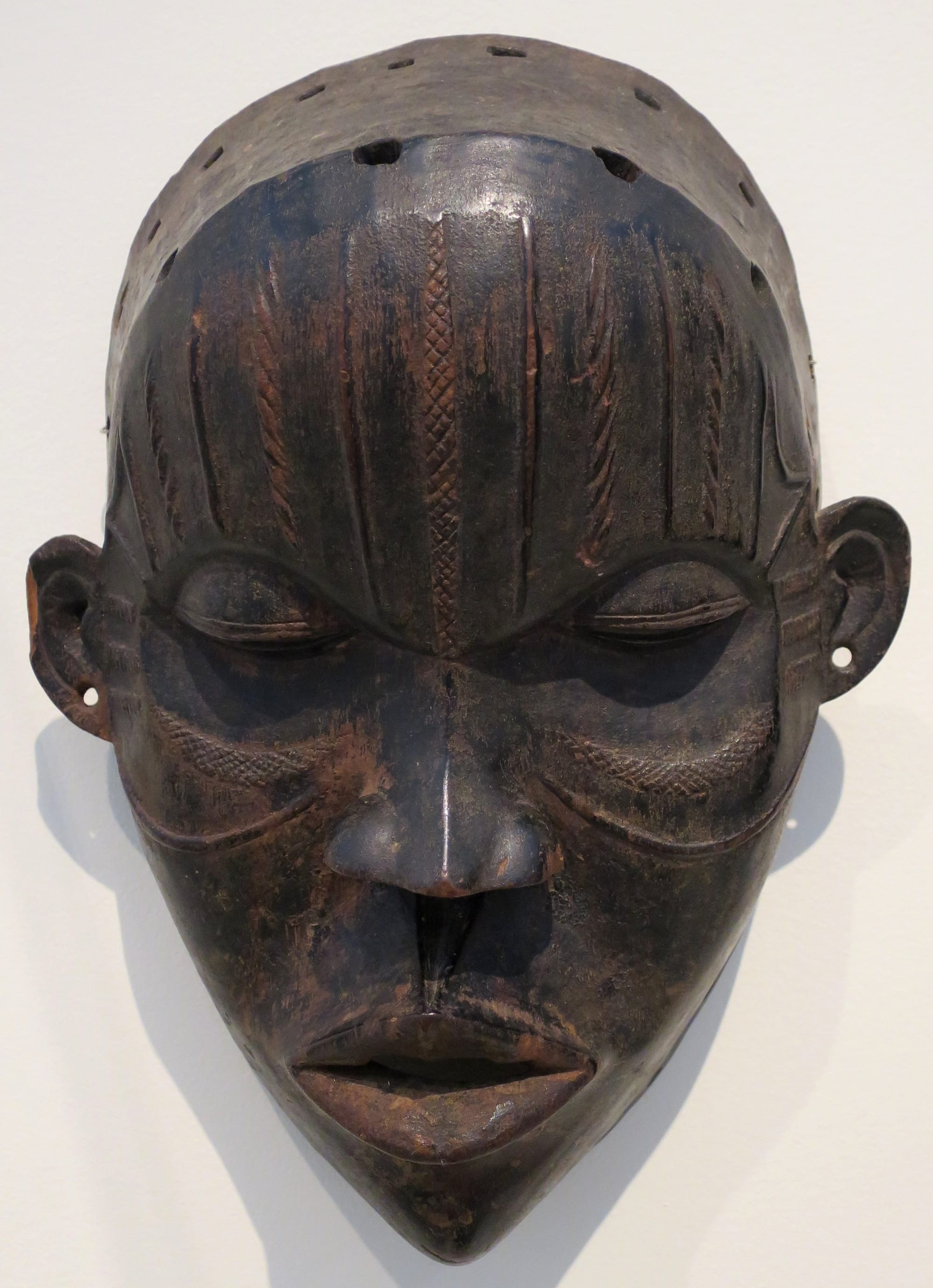 A dark carved wooden face mask with narrow slit eyes, full parted lips, small ears and incised vertical lines across the forehead, displayed against a white wall