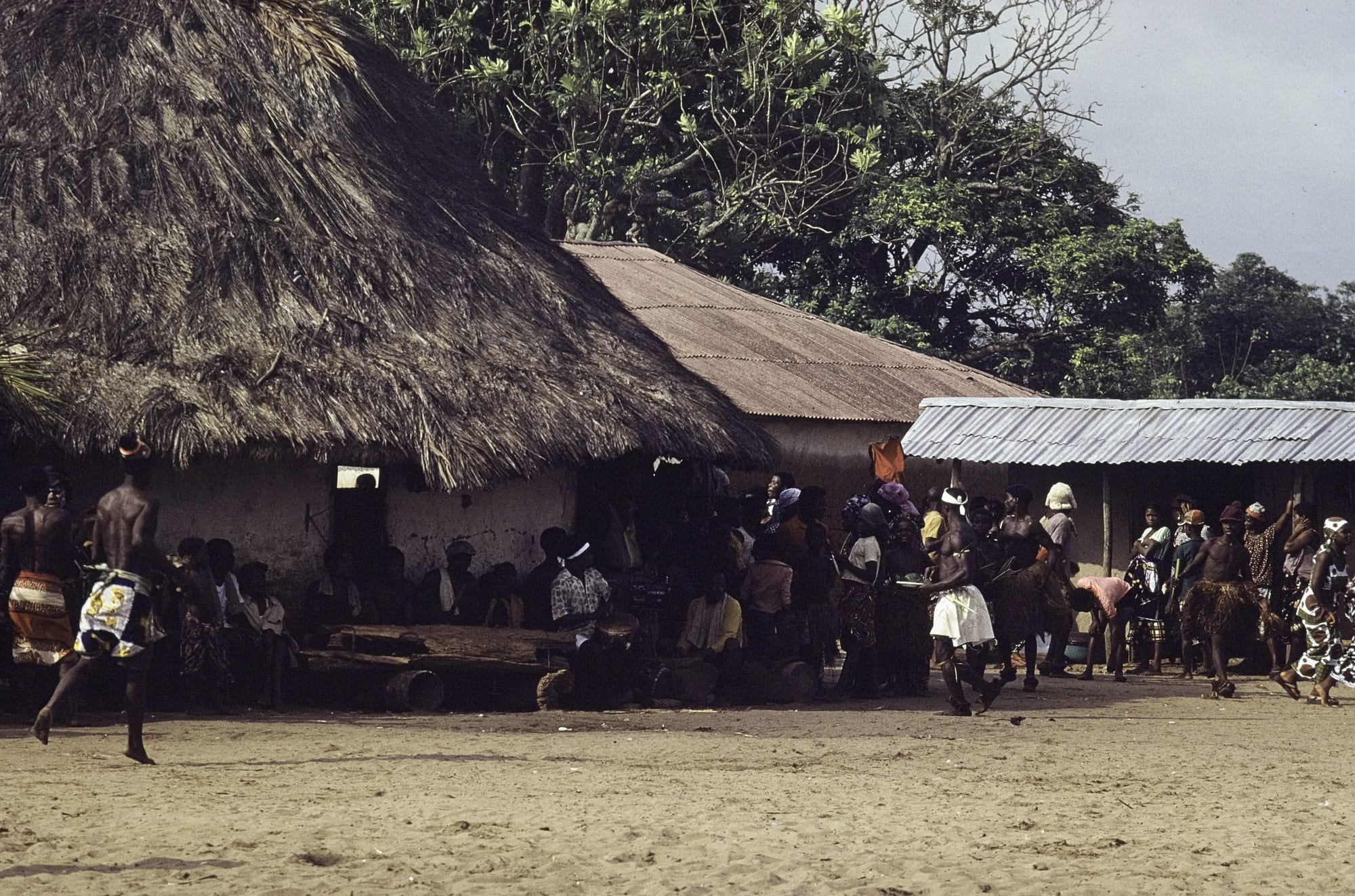 Drummers in the shade of a large thatched roof play a long horizontal log drum while dancers in straw skirts and printed cloth move across a sandy village square, watched by a crowd