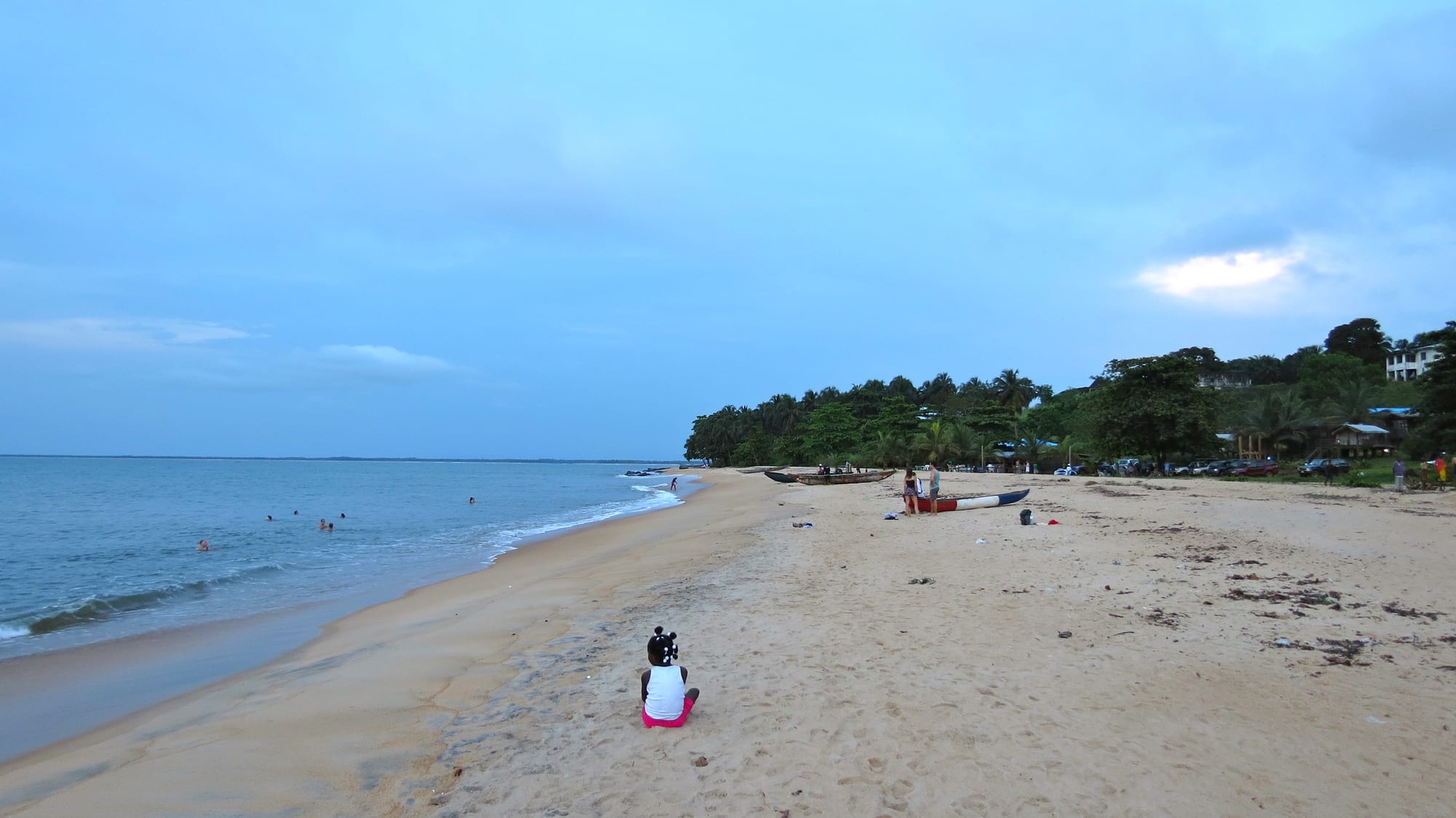 A wide sandy beach at dusk with fishing canoes, palm trees, and people gathered by the water