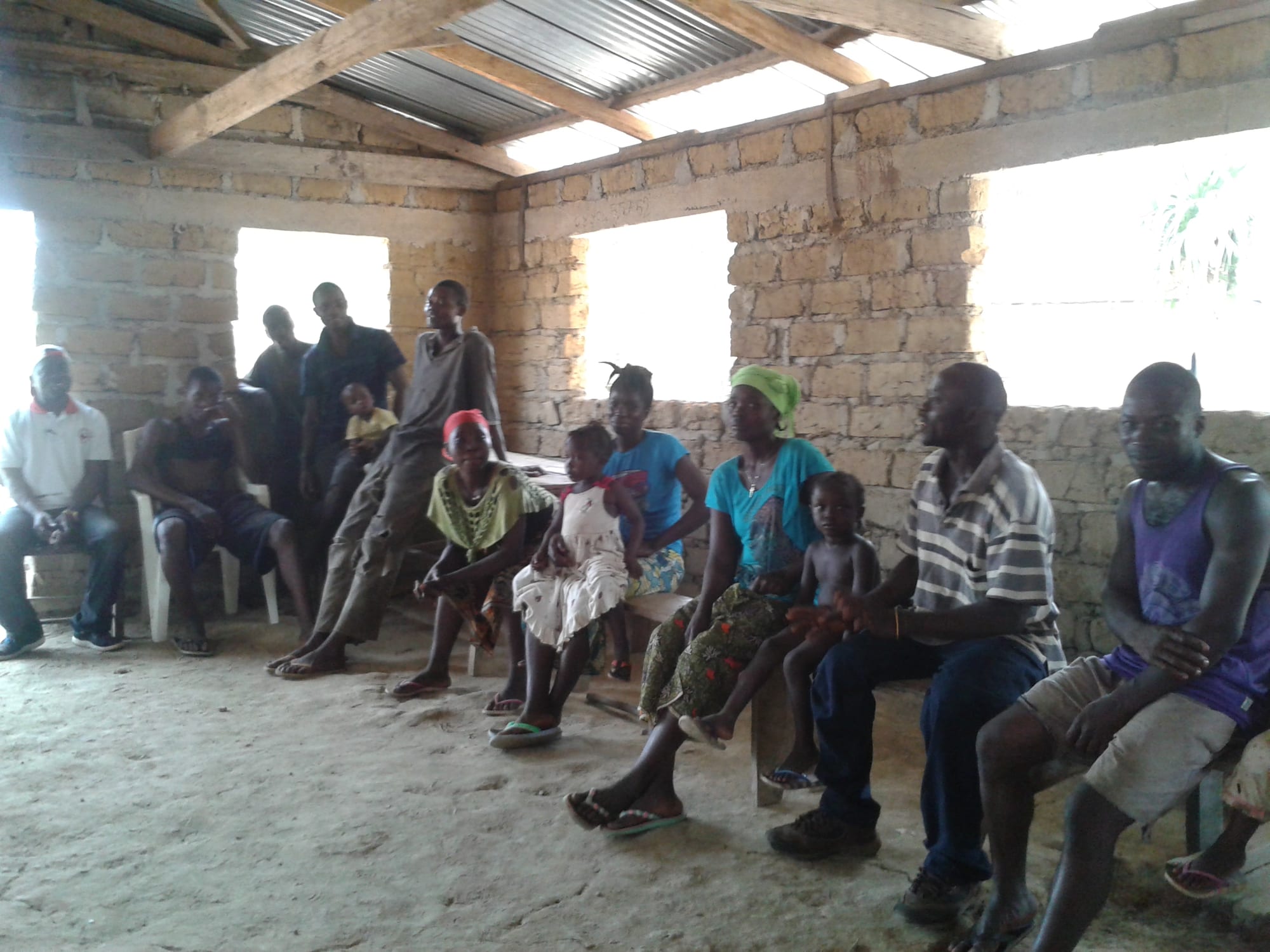 A large group of adults and children of different ages sitting and standing together inside a partly built brick building with a corrugated roof
