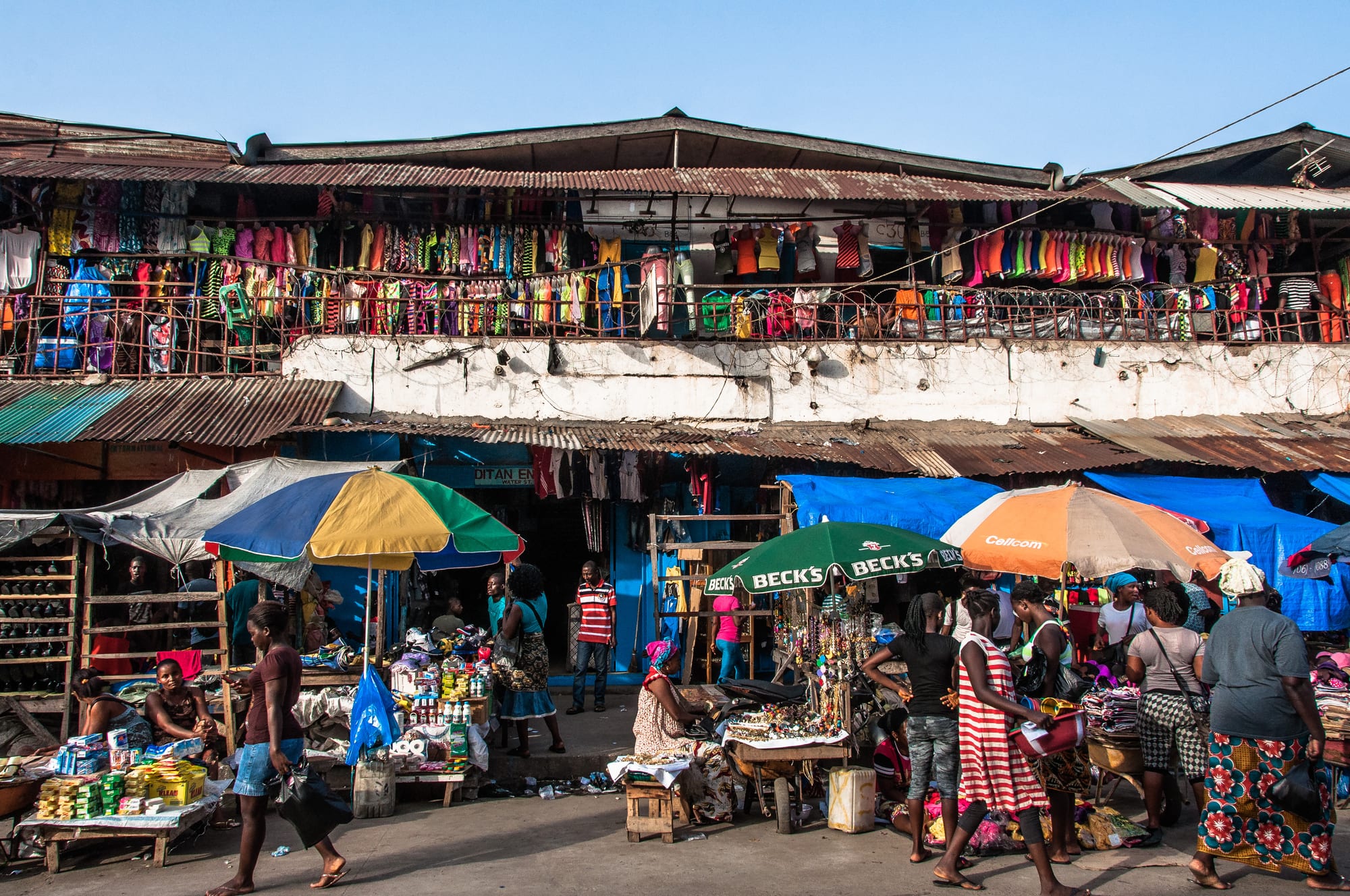 A row of market stalls under colourful umbrellas, with a second-storey balcony hung with clothing for sale and shoppers moving through the street below
