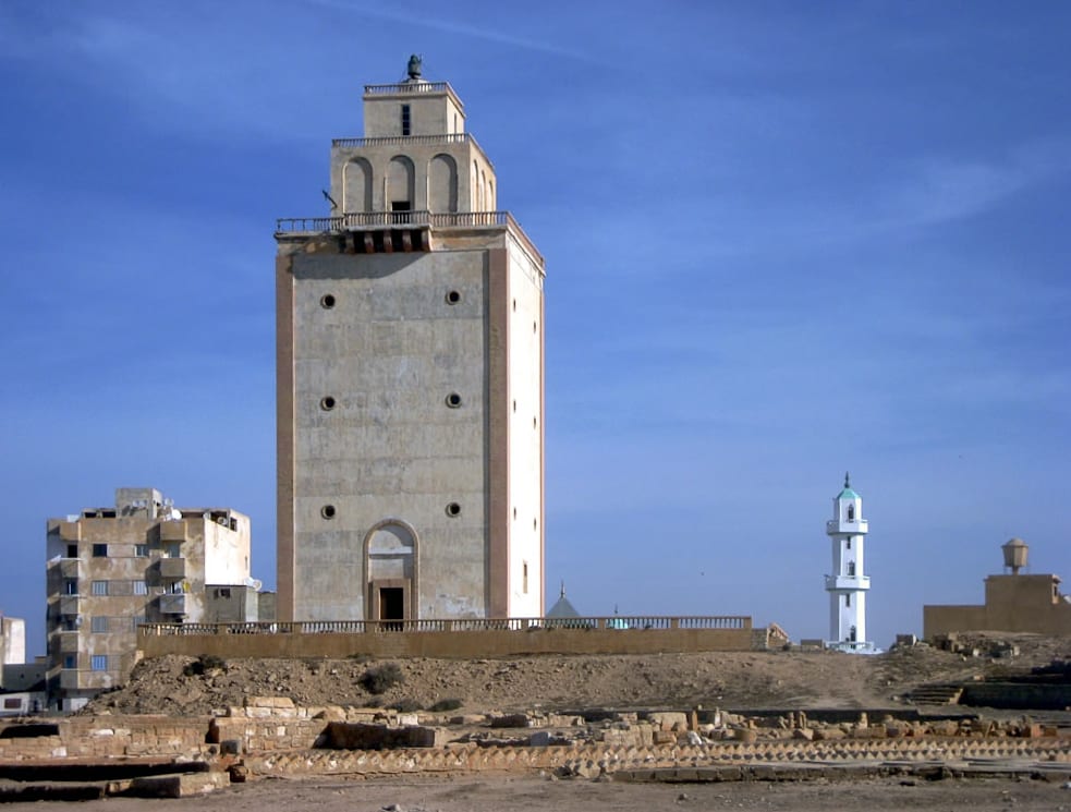 A tall white Italian-era lighthouse on a Mediterranean waterfront