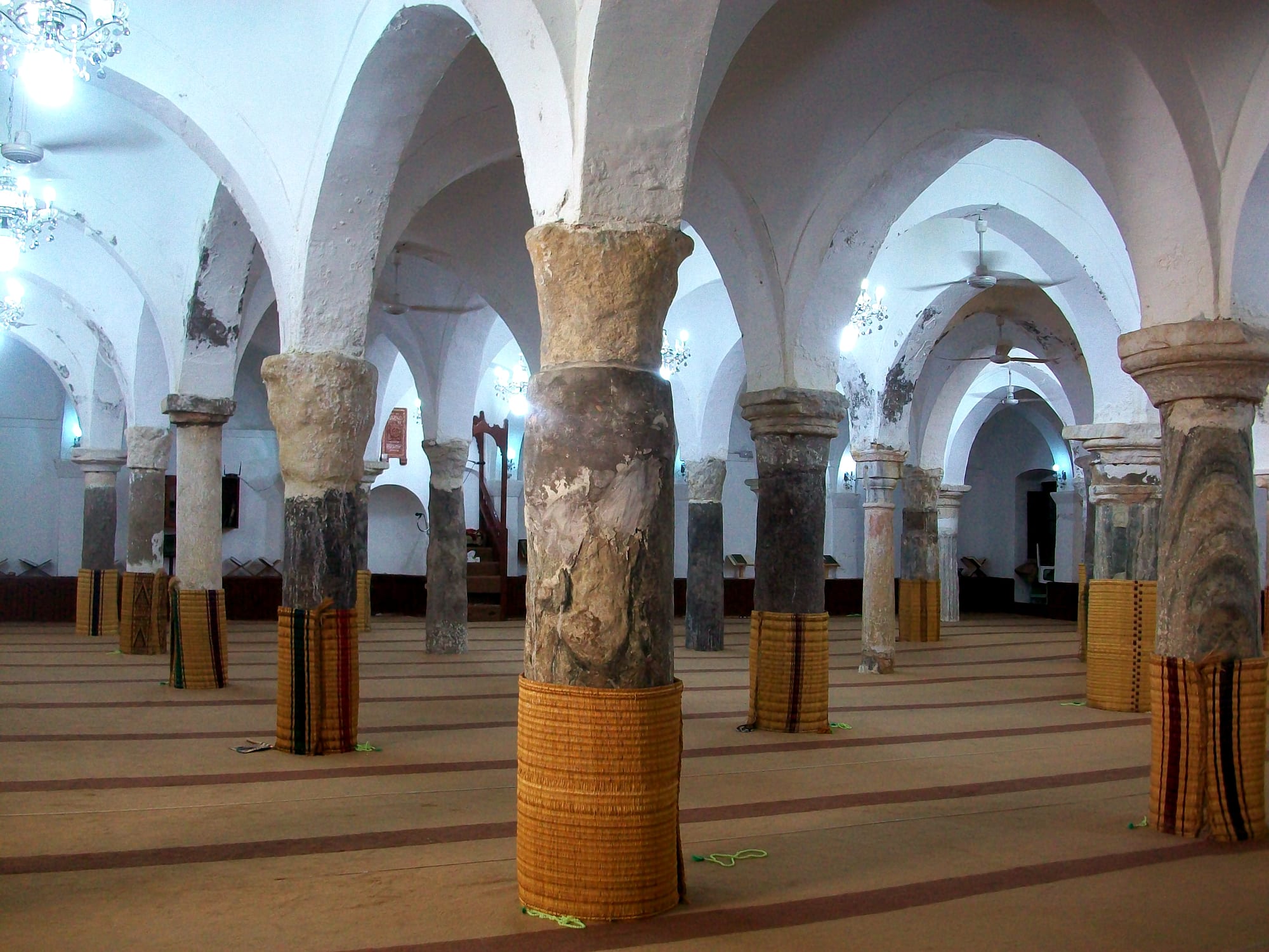 Rows of stone columns of different colours, their lower parts wrapped in woven matting, support whitewashed arches over a carpeted prayer hall