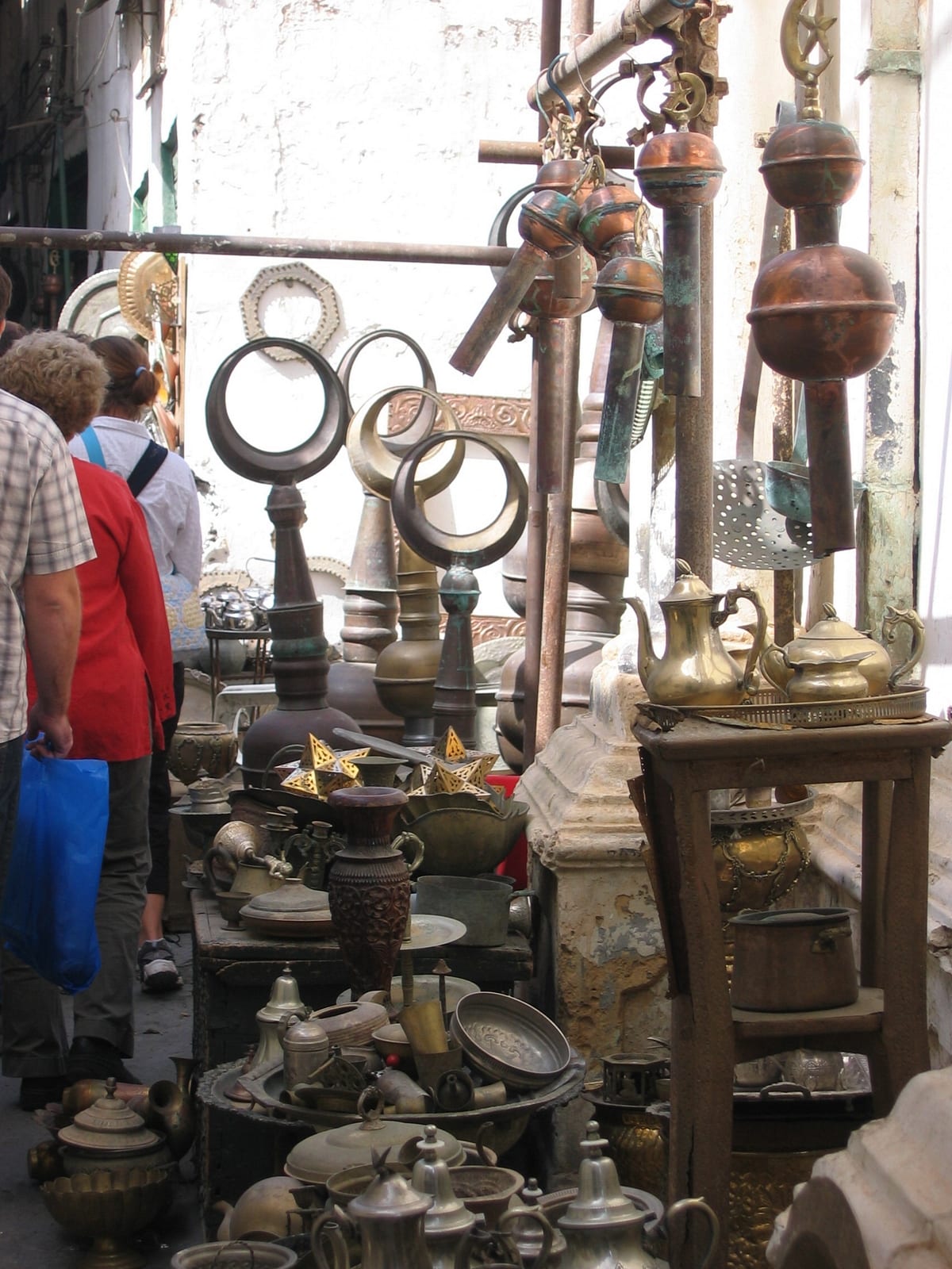 A narrow market lane crowded with brass teapots, copper jugs, trays, star-shaped lanterns and tall metal crescent finials, with shoppers walking past