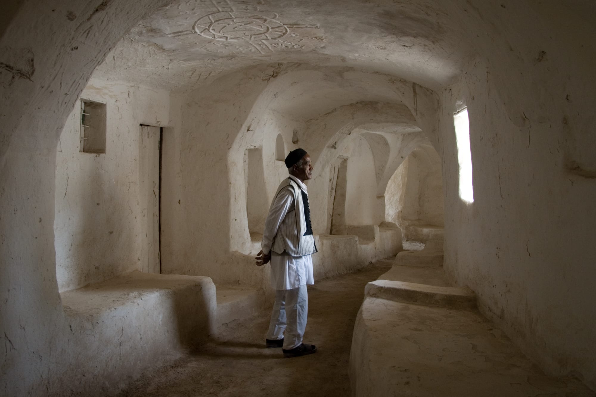 A man in a white tunic, striped waistcoat and dark cap stands in a whitewashed vaulted passage lined with built-in benches, lit by a small window