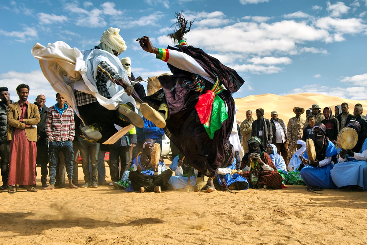 A dancer in flowing white and dark robes leaps high above the sand while women with frame drums sit in a circle and spectators watch, with pale dunes behind