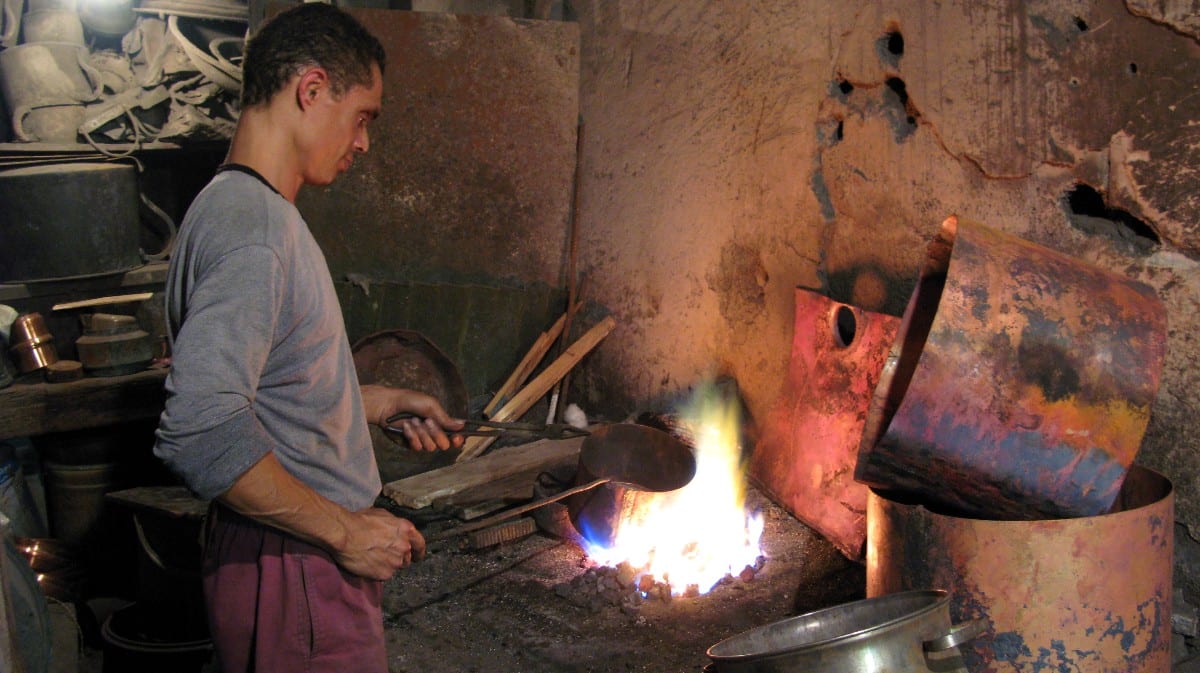 A metalworker in a workshop heating metal over an open flame, surrounded by copper and tin vessels