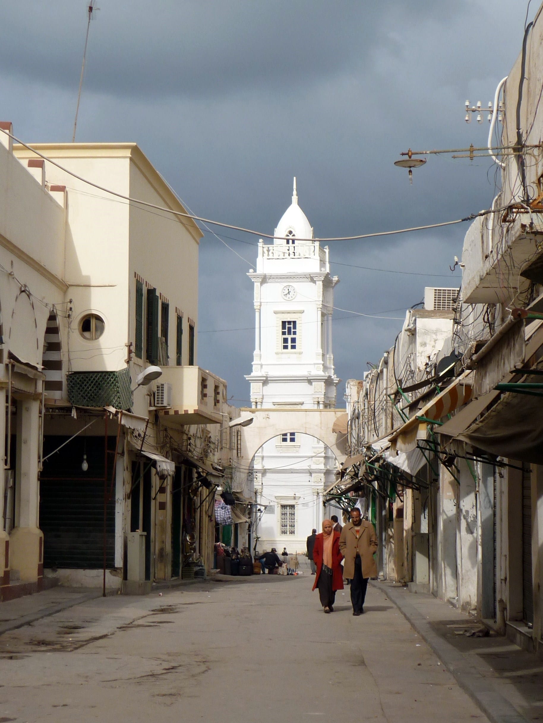 A couple walking down a narrow street of shuttered shops in Tripoli's old town, with a white Ottoman-era clock tower rising ahead of them under a stormy sky