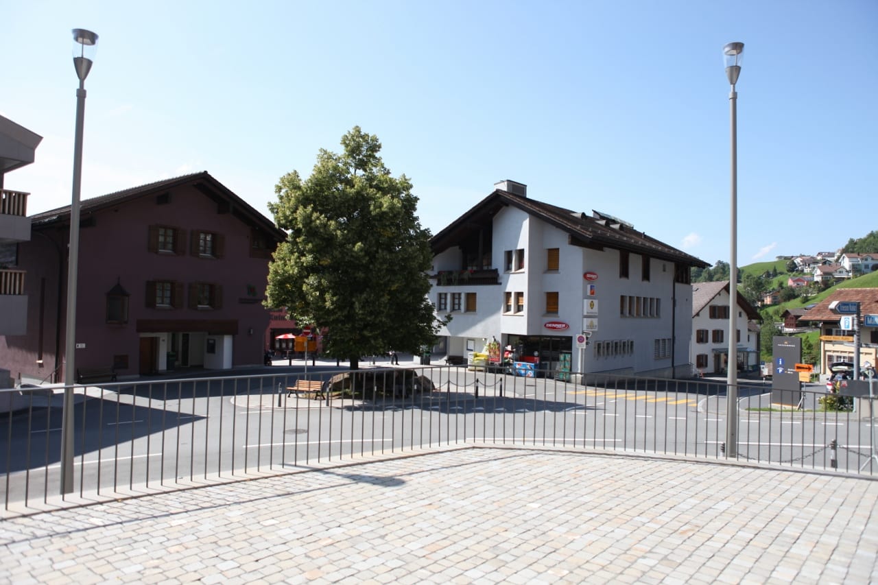 A small village square in Triesenberg with traditional chalet-style houses, a corner shop, and green hillsides behind, on a sunny day