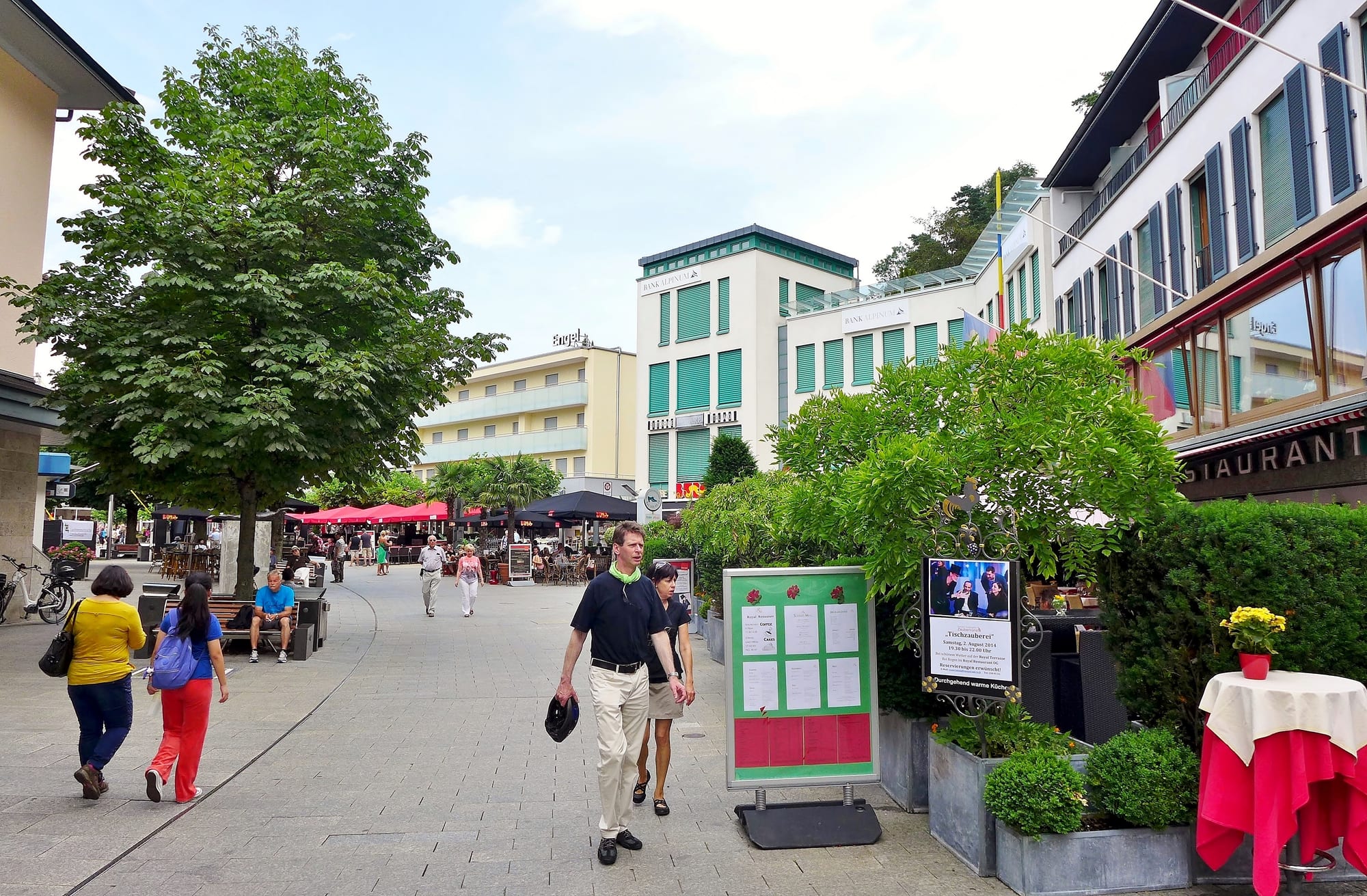 People walking and sitting at outdoor café tables on a tree-lined pedestrian street in Vaduz, with low modern buildings on either side