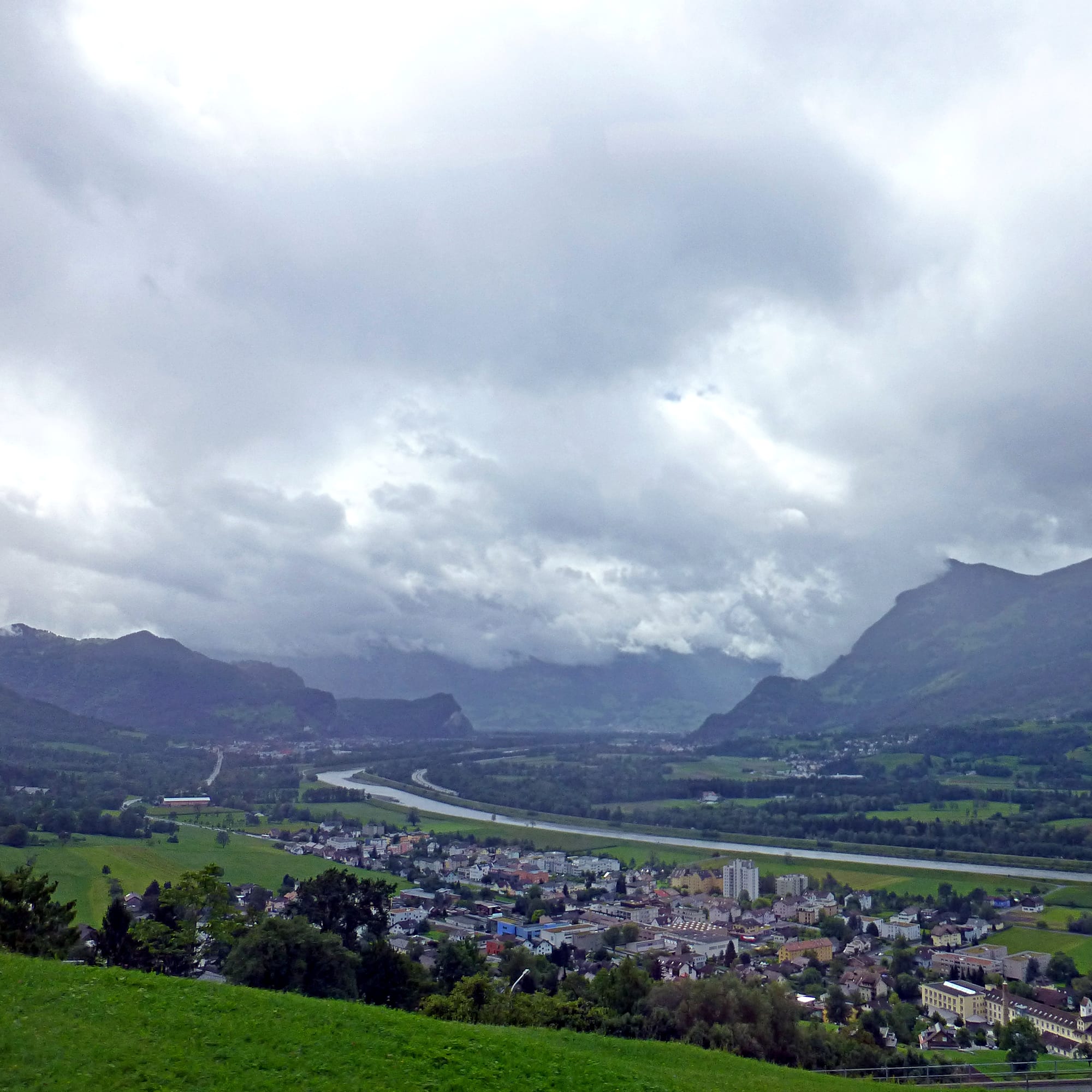 A wide green valley of farmland and villages seen from a mountainside, mountains beyond