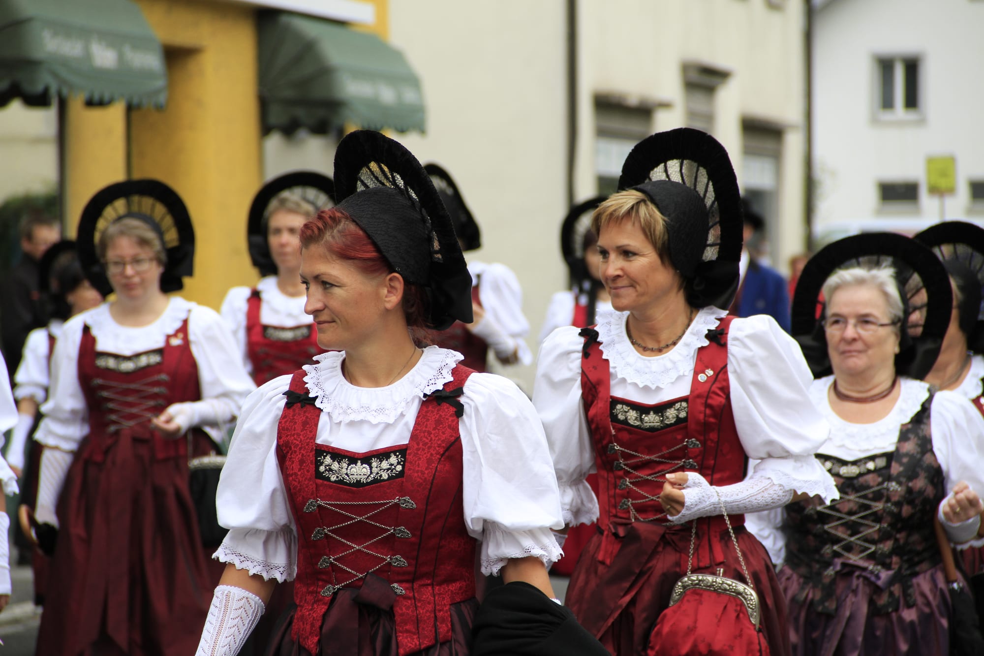 Women in traditional costume with red laced bodices, white puffed sleeves and tall black fan-shaped bonnets walking in a street parade