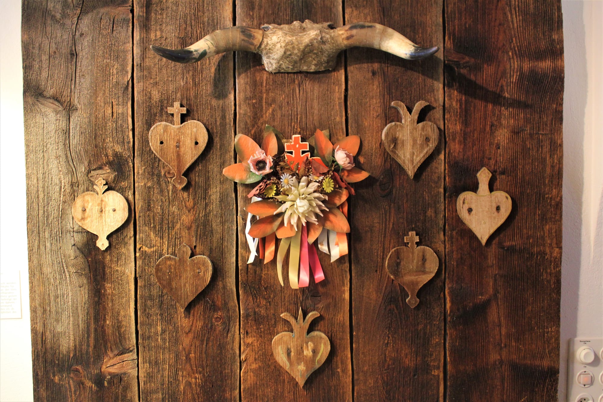 Carved wooden hearts of several shapes, some topped with crosses, fixed to weathered boards around a spray of flowers and ribbons, beneath a pair of cattle horns