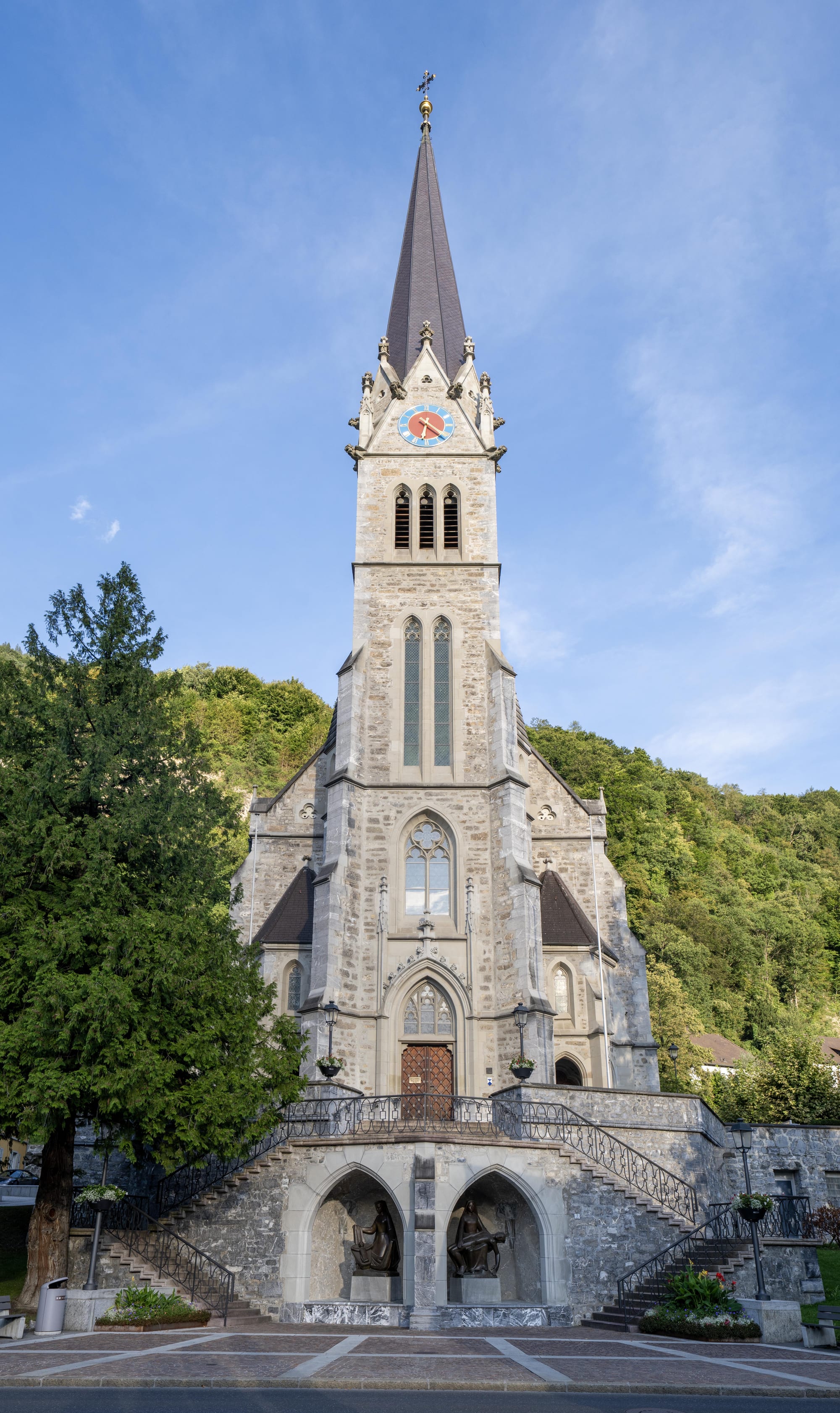 A grey stone neo-Gothic church with a tall pointed spire and clock, twin flights of steps leading up to the entrance, with wooded hillside behind