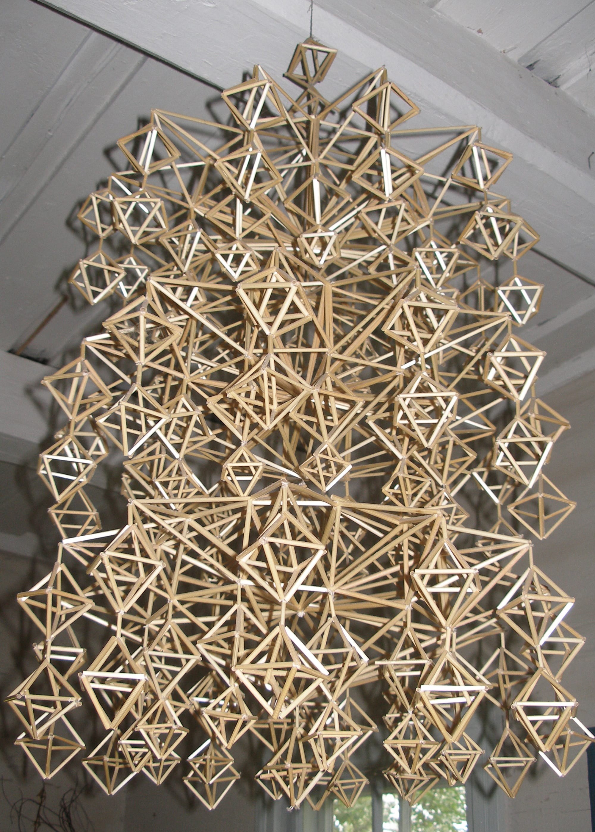 A large hanging ornament made of hundreds of straw stalks joined into interlocking diamond and pyramid shapes, suspended from a white wooden ceiling