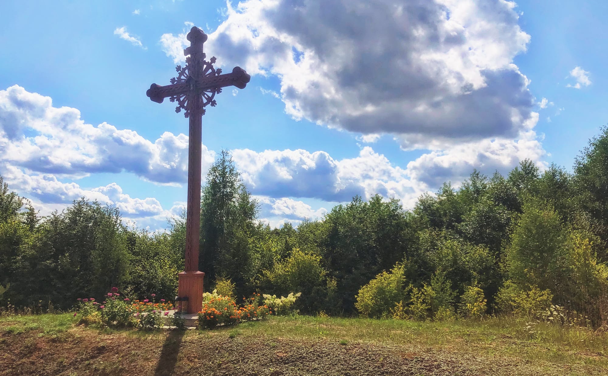 A tall carved wooden cross with an ornate sunburst at its centre stands on a small plinth among flower beds, with green woodland and a blue sky full of white clouds behind