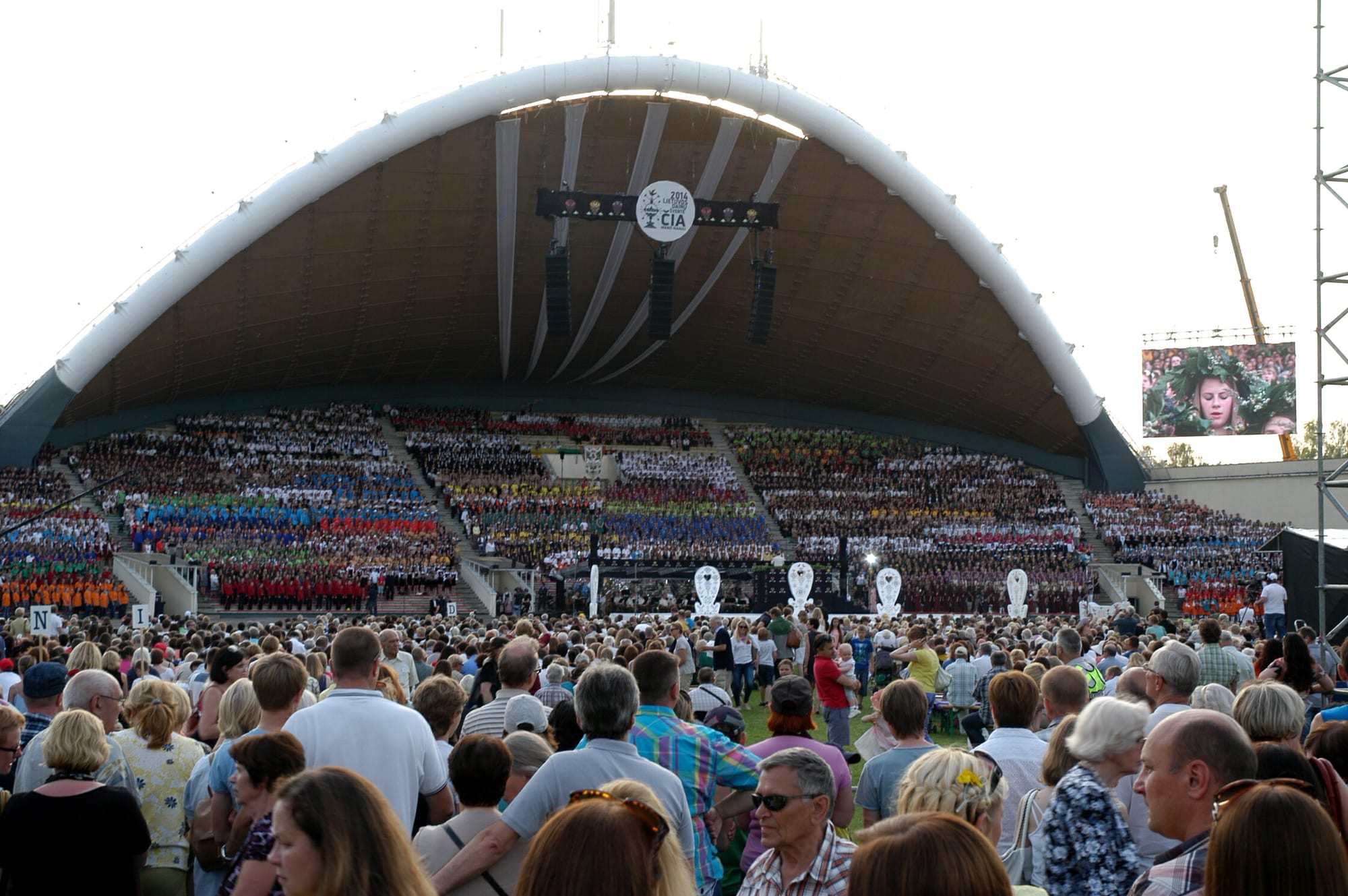 Thousands of singers in coloured costumes filling the tiered stage beneath a large white arched shell, with a big audience standing in front