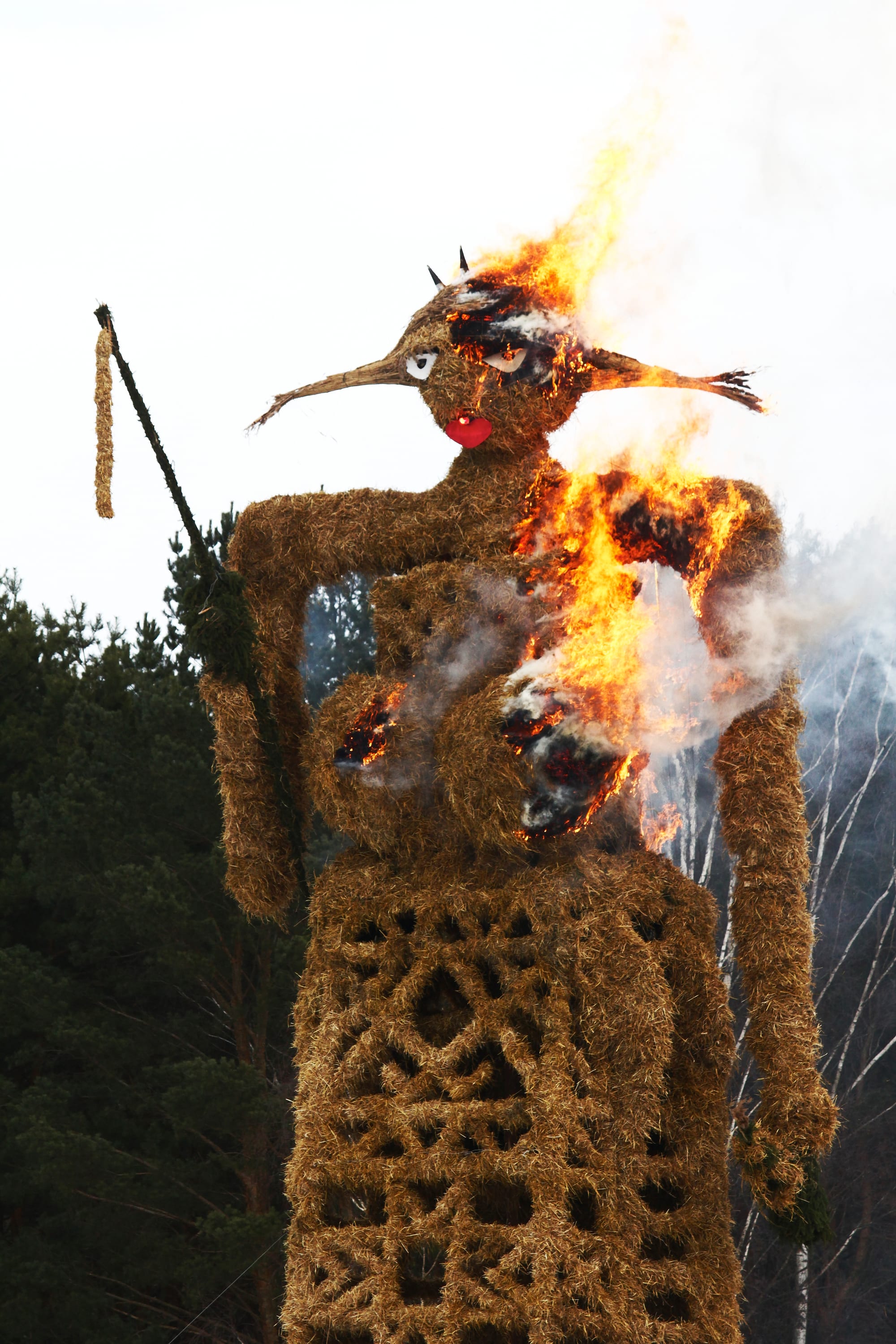 A tall woven straw figure with a long beak-like nose, red lips and a patterned straw skirt, with flames starting to climb its body against pine trees