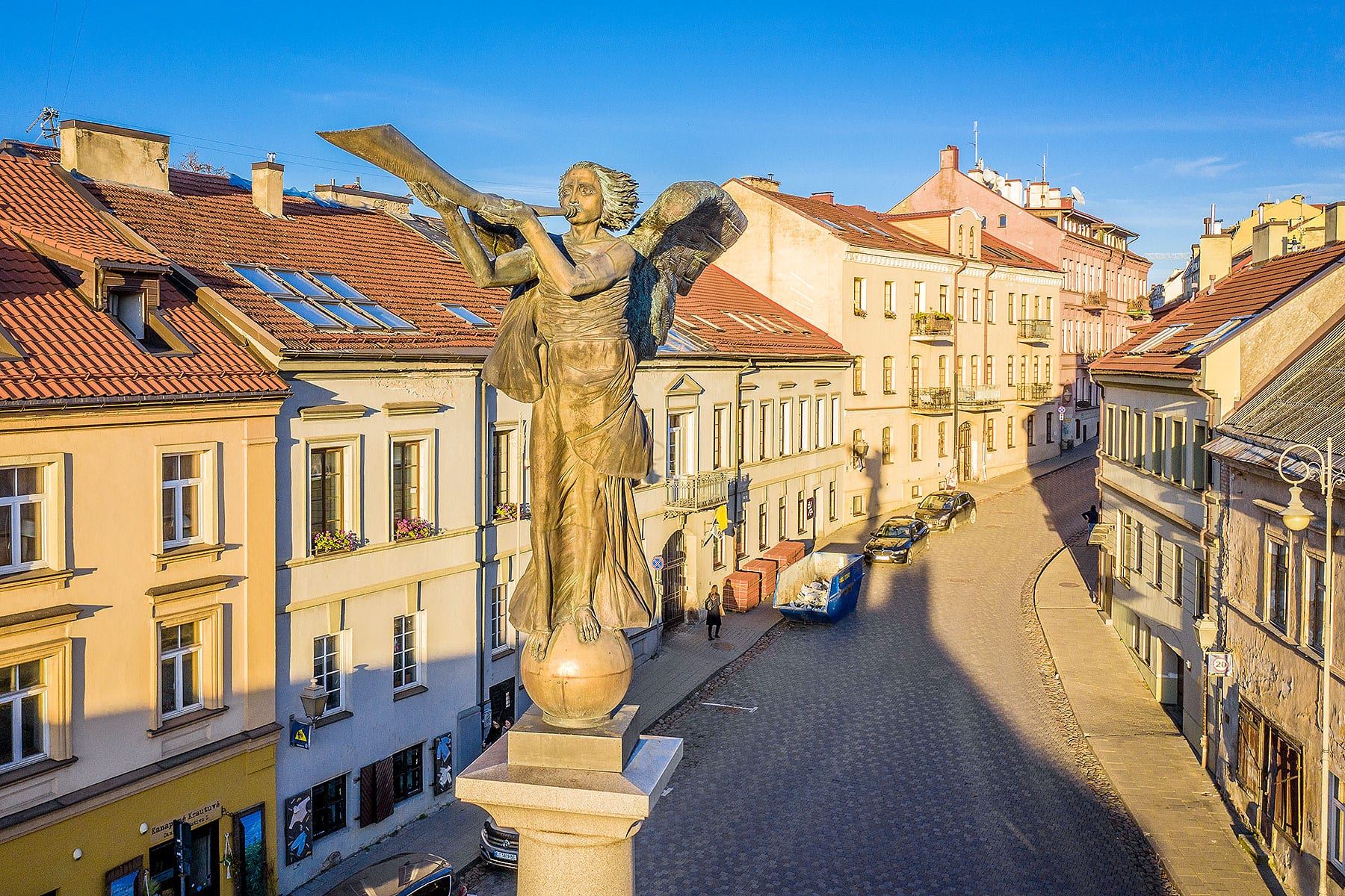 A bronze angel statue on a column above a cobbled street of colourful old buildings