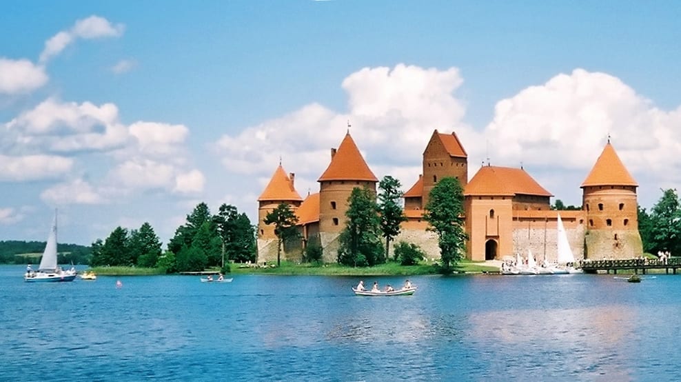 A red-brick medieval castle on a small island, connected by wooden bridges, surrounded by a lake