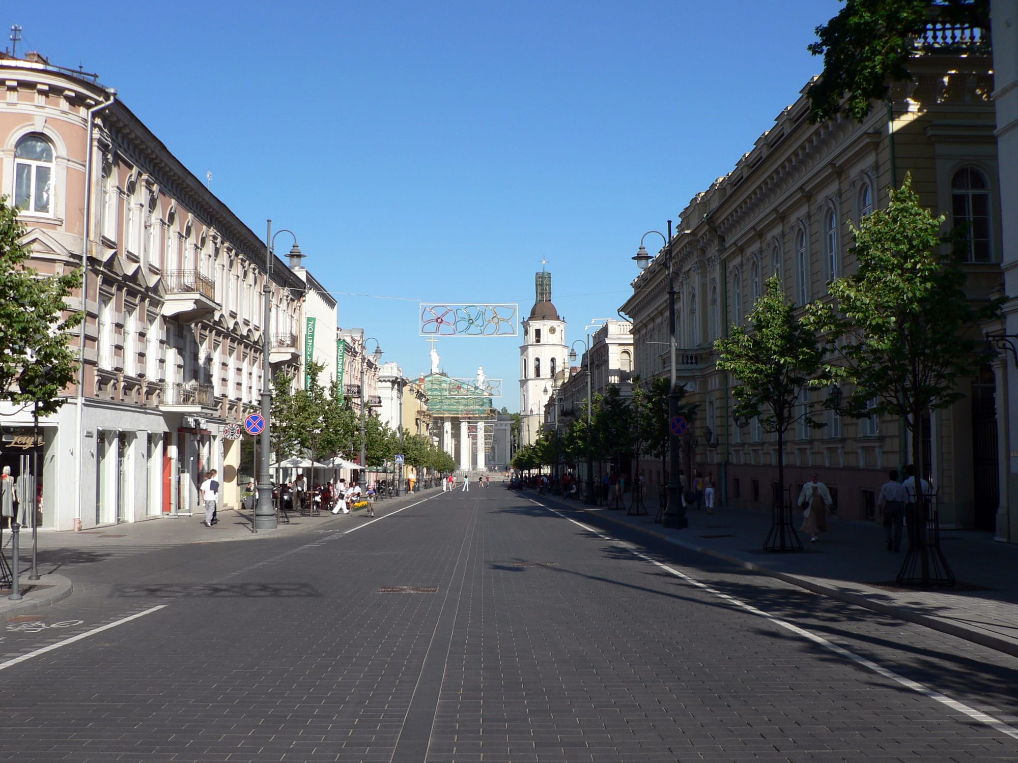 A wide pedestrianised avenue lined with pastel-coloured buildings and trees, with people walking and a cathedral belfry in the distance