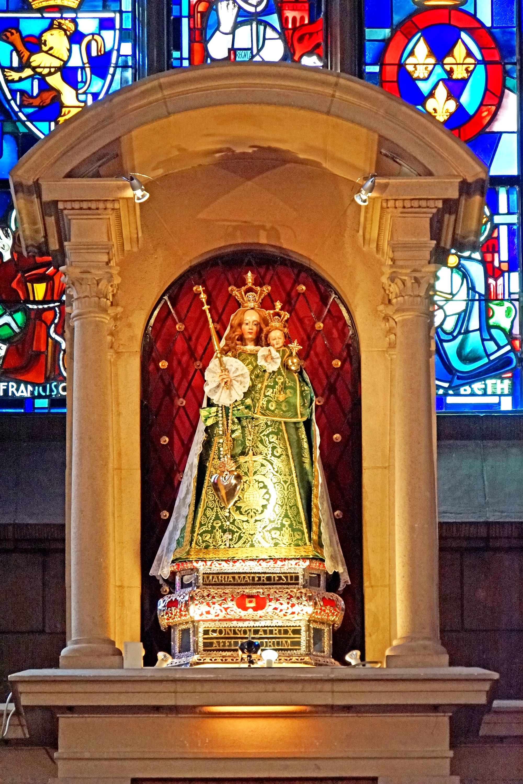 A crowned statue of the Virgin Mary holding the infant Jesus, dressed in an embroidered green robe with a lace veil, standing in a lit stone niche in front of stained-glass windows