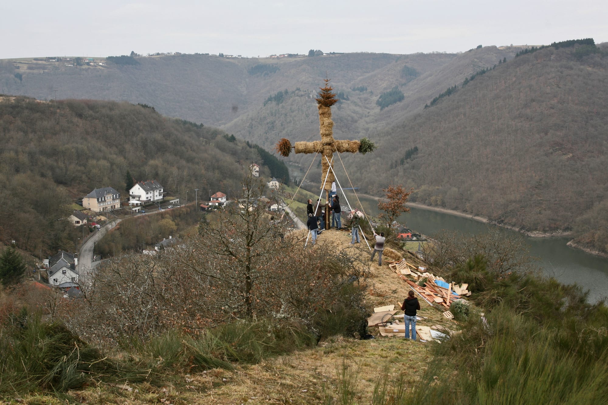 A group of people on a hilltop raising a tall cross made of bundled straw with evergreen tips, held by guy ropes, above a village and a winding river in a wooded valley