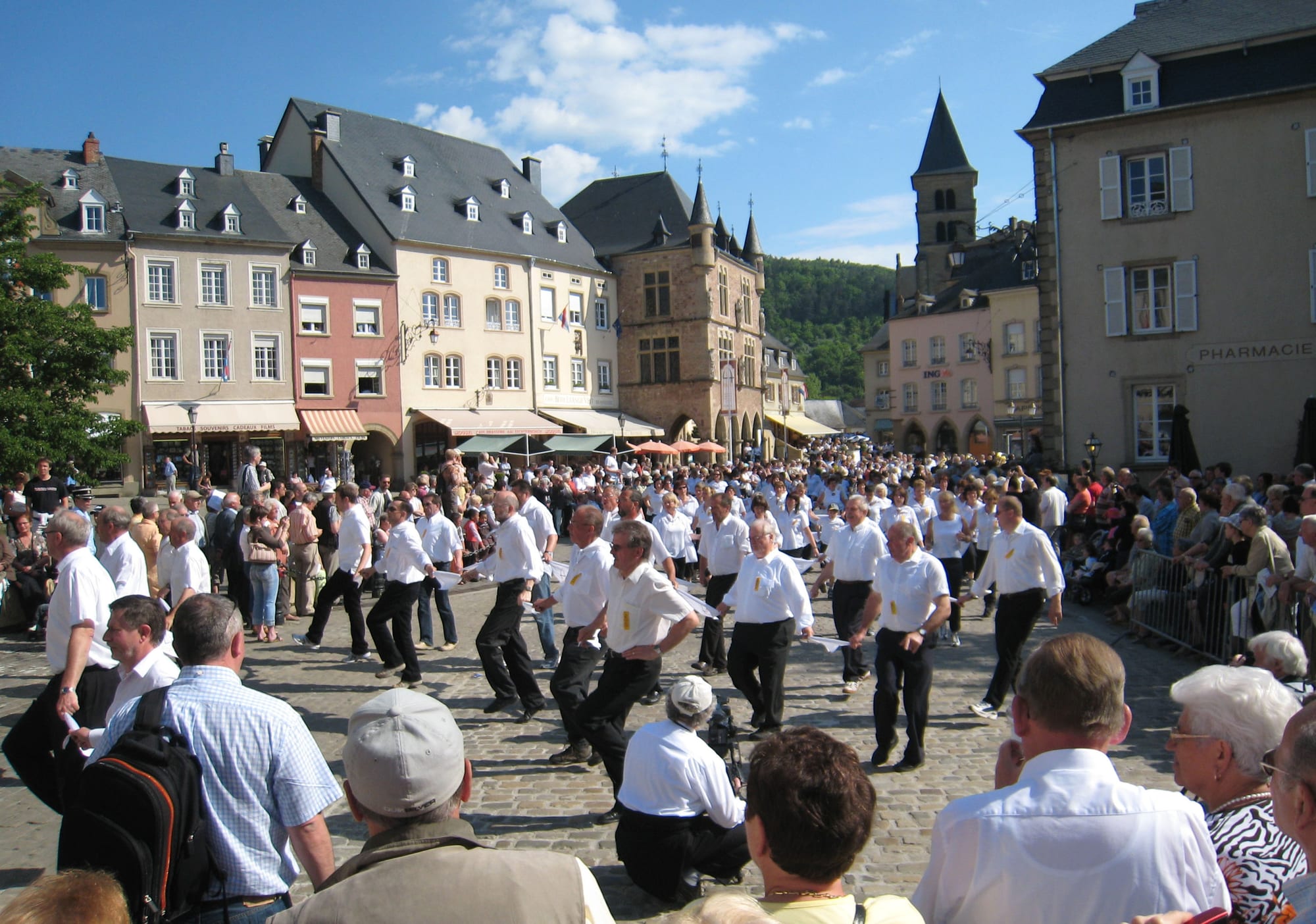 Rows of men in white shirts and dark trousers, linked by white handkerchiefs, hopping across a cobbled square lined with tall old houses while crowds watch