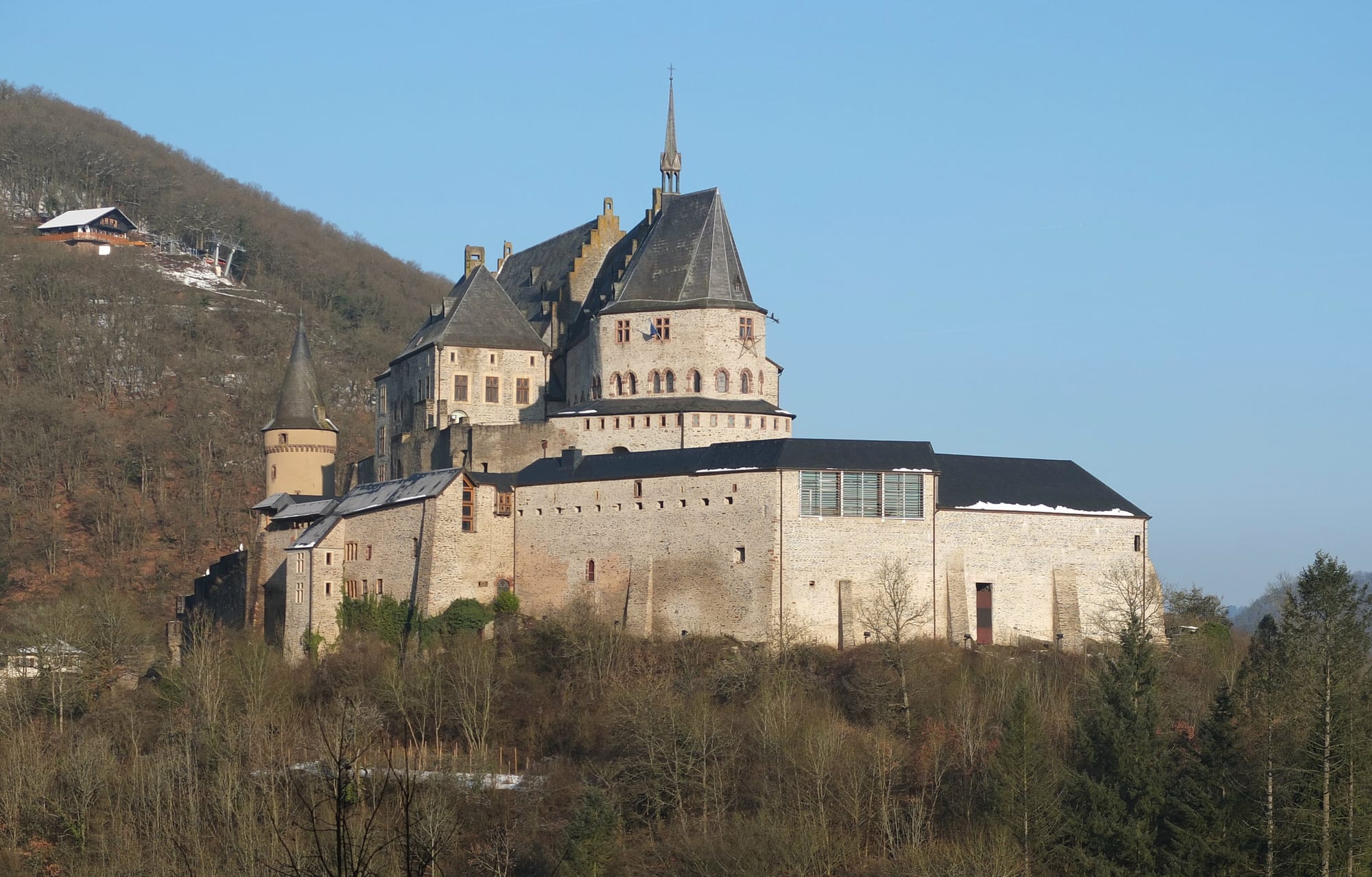 A large medieval castle with round towers crowning a hilltop above a small river town