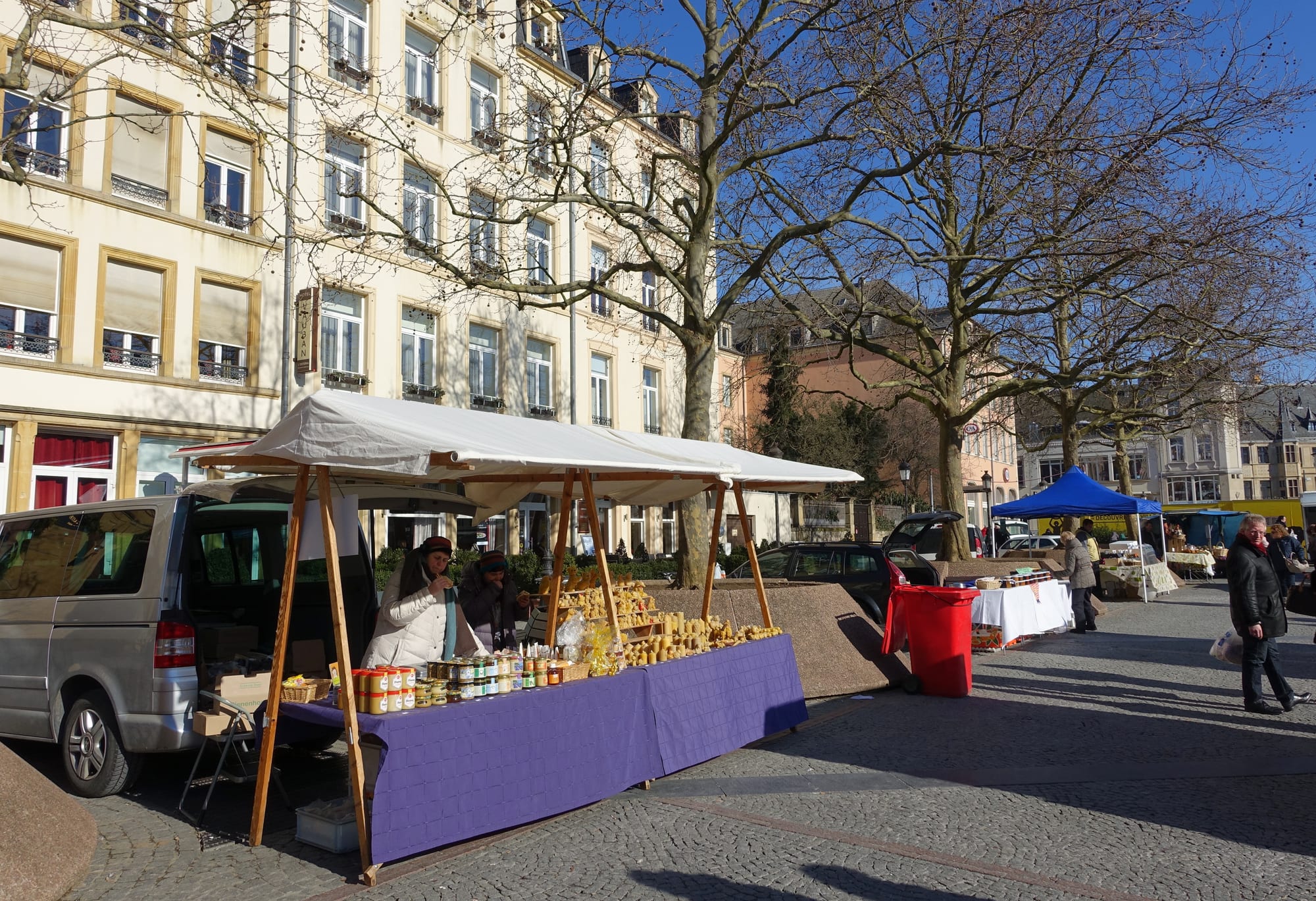 An outdoor market with striped awnings and stalls selling produce, set up in a paved city square surrounded by buildings
