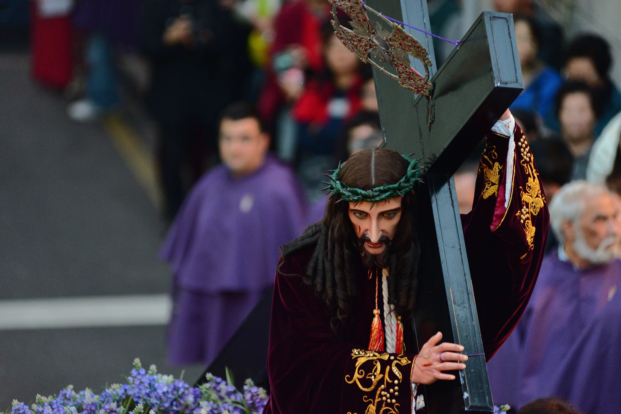 A carved statue of Christ in a crown of thorns and a dark red and gold embroidered robe, carrying a black cross, with purple-robed participants and onlookers blurred behind