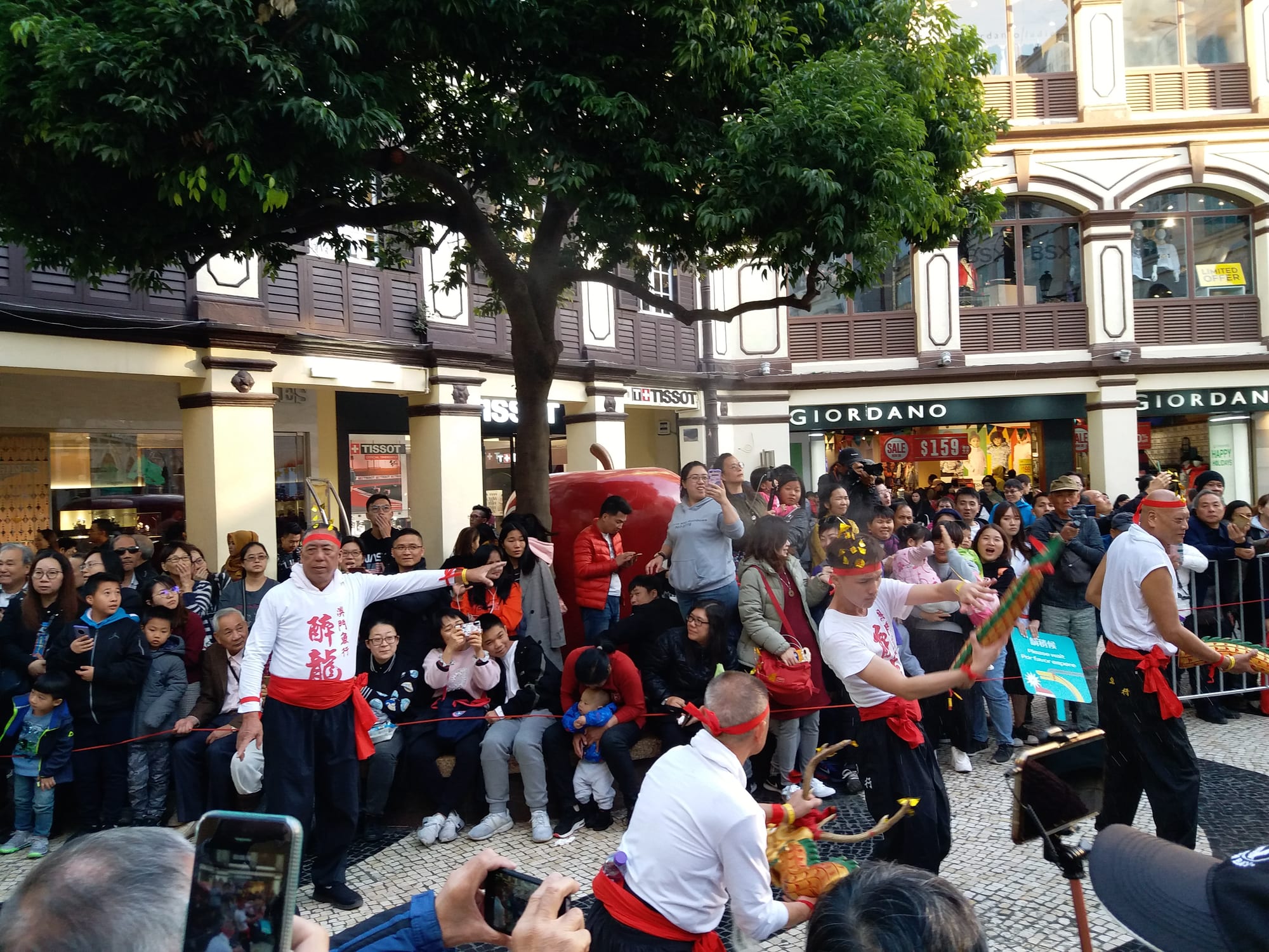 Performers in white tops with red sashes and headbands dancing with carved wooden dragon heads and tails on a paved square, watched by a large crowd in front of shopfronts