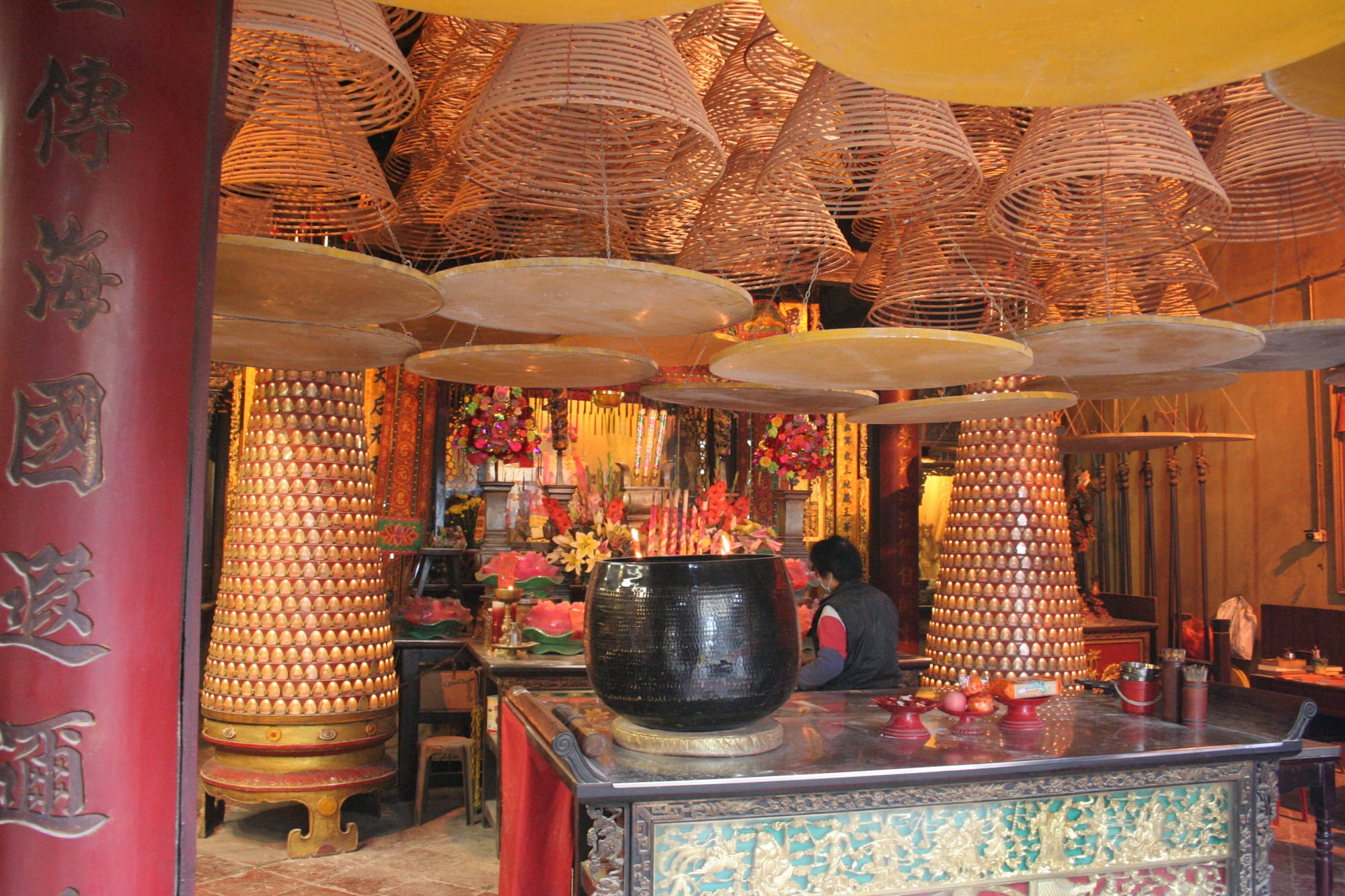 Dozens of large cone-shaped incense coils hanging from the ceiling of a temple hall above a black iron incense burner, an altar with flowers and towers of small offerings