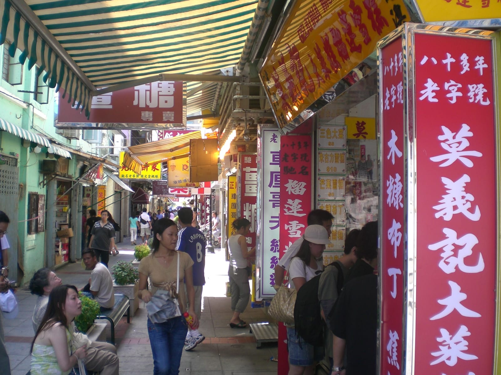 A narrow pedestrian street in Taipa Village crowded with shoppers under striped awnings and large Chinese shop signs