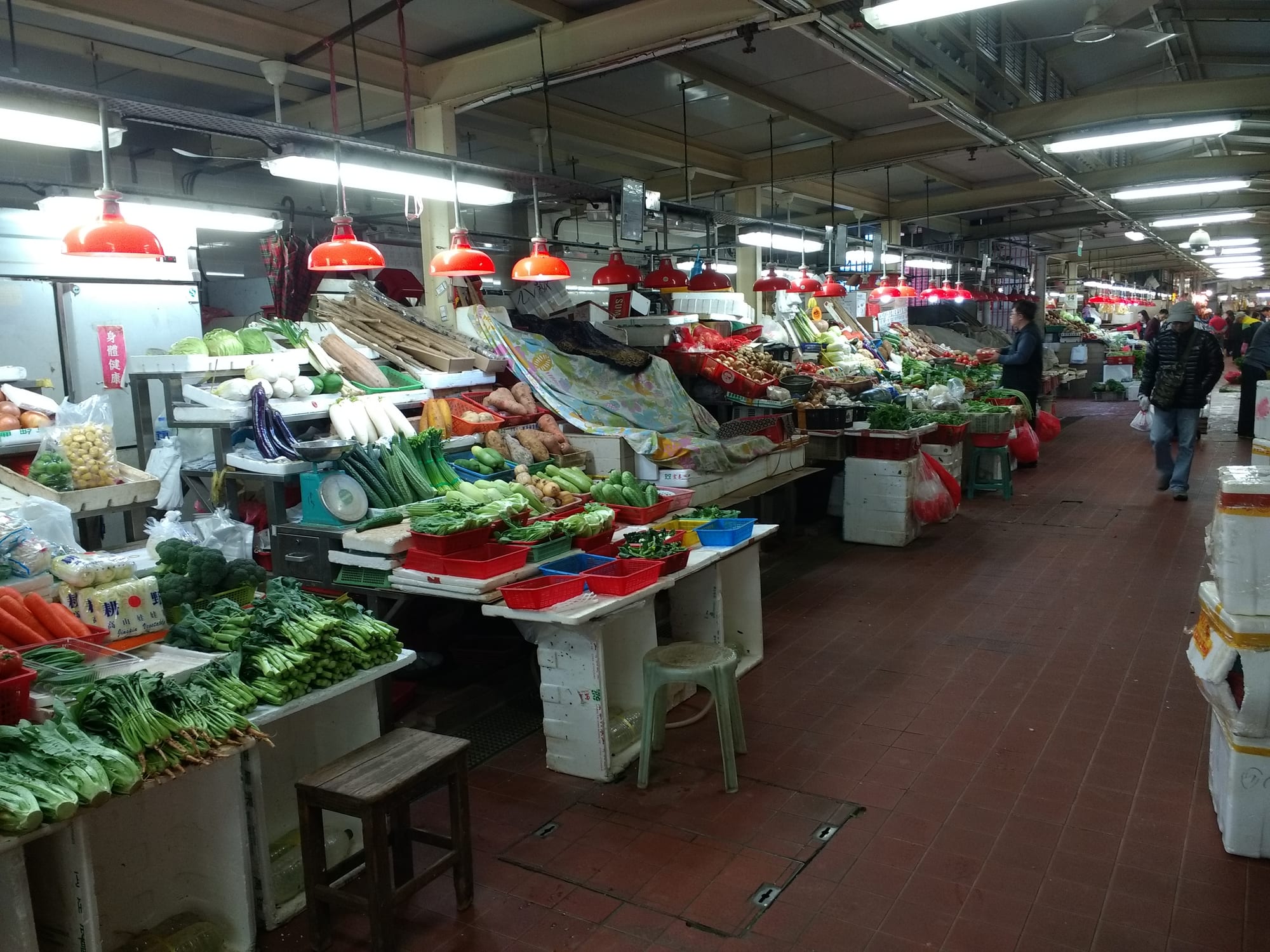 Rows of market stalls piled with fresh vegetables under red lamp shades inside a covered market hall
